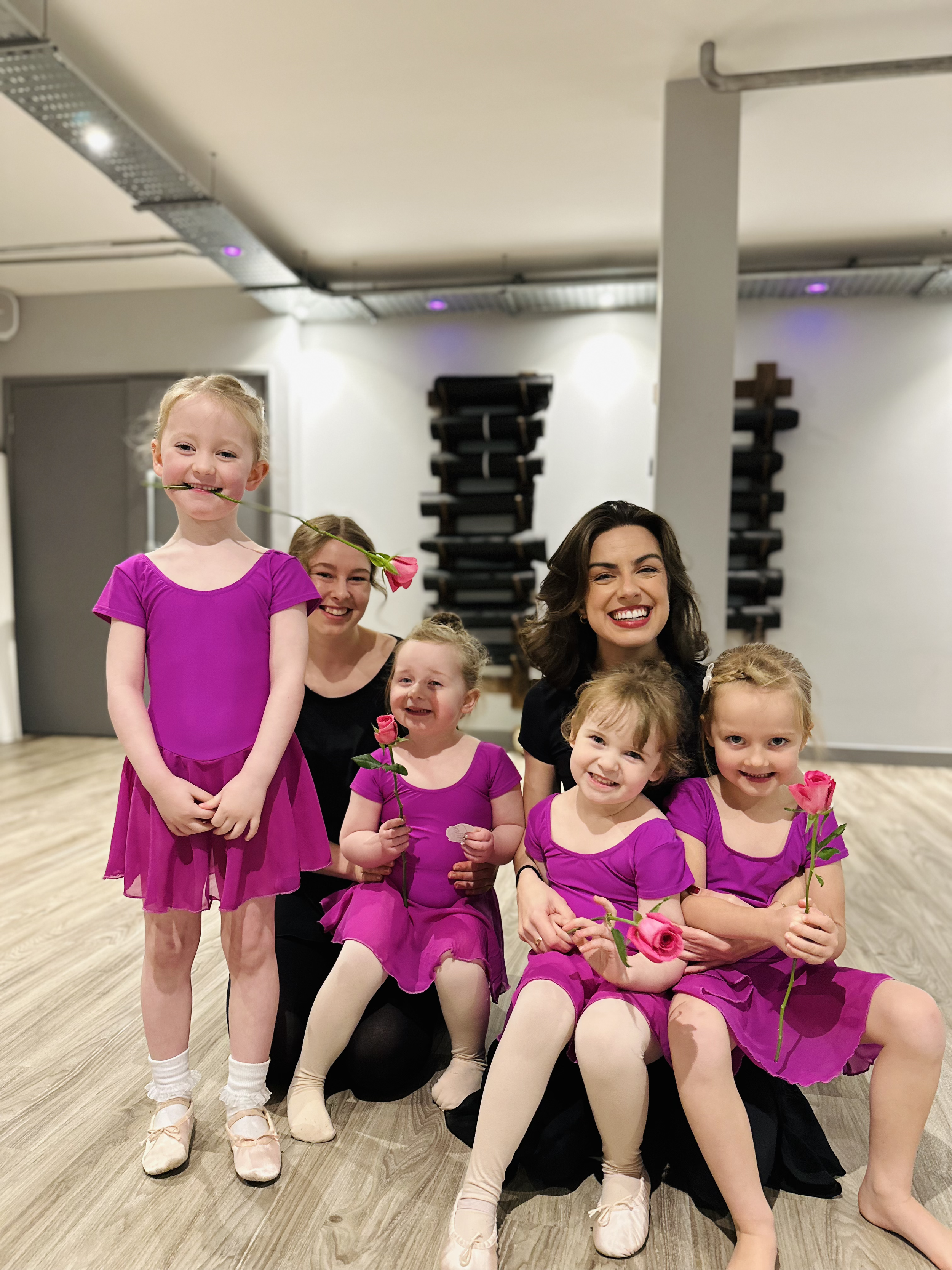 Five young girls in purple dance costumes with white tights and ballet slippers, along with two women, one in a black dress and another in a black shirt, smiling and holding pink roses, in a dance studio.