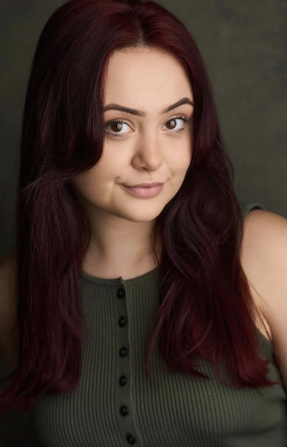 A young woman with red hair, smiling and wearing a dark green sweater, sitting against a brown background.