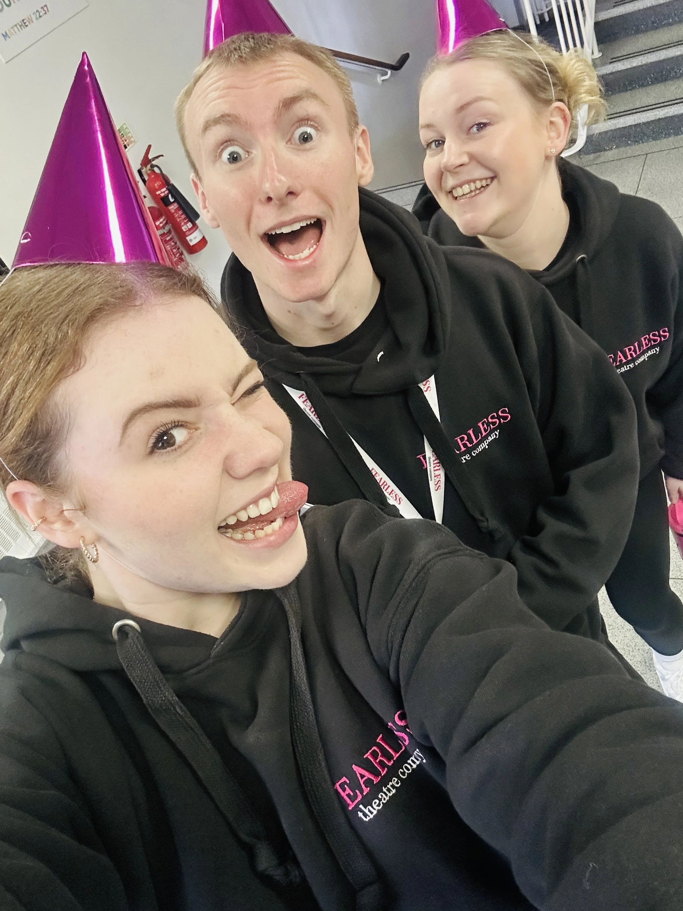 Four young people wearing black hoodies and pink party hats taking a selfie. Two are making funny faces, one winking and sticking out her tongue, all smiling.