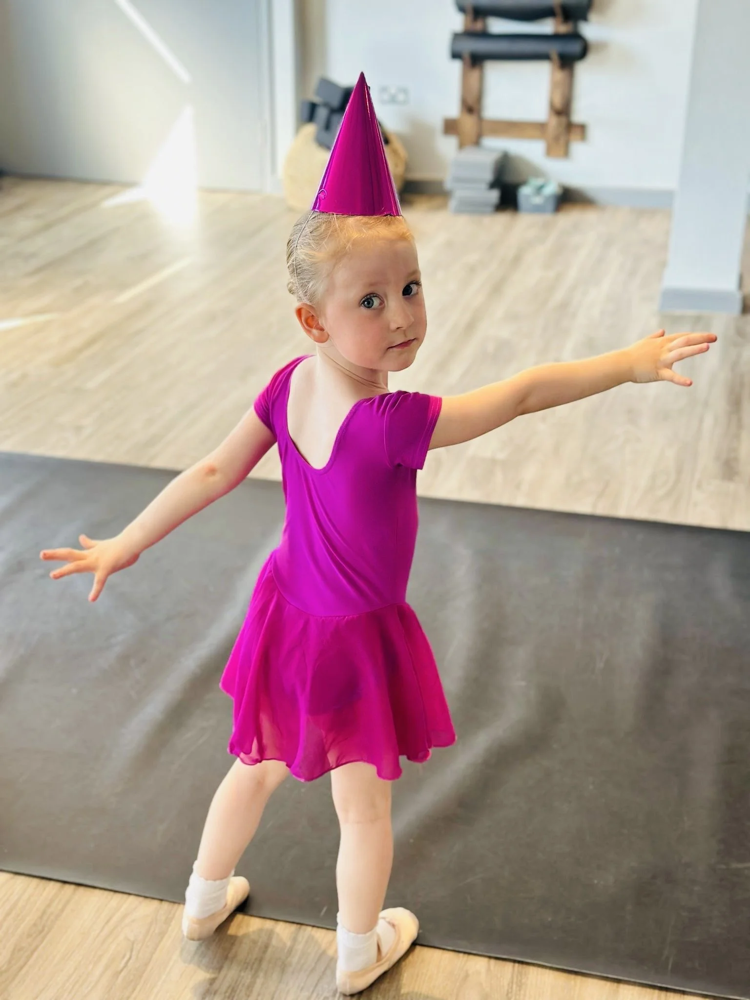 Young girl dressed in a bright pink dance costume and matching party hat performing a dance pose in a studio.