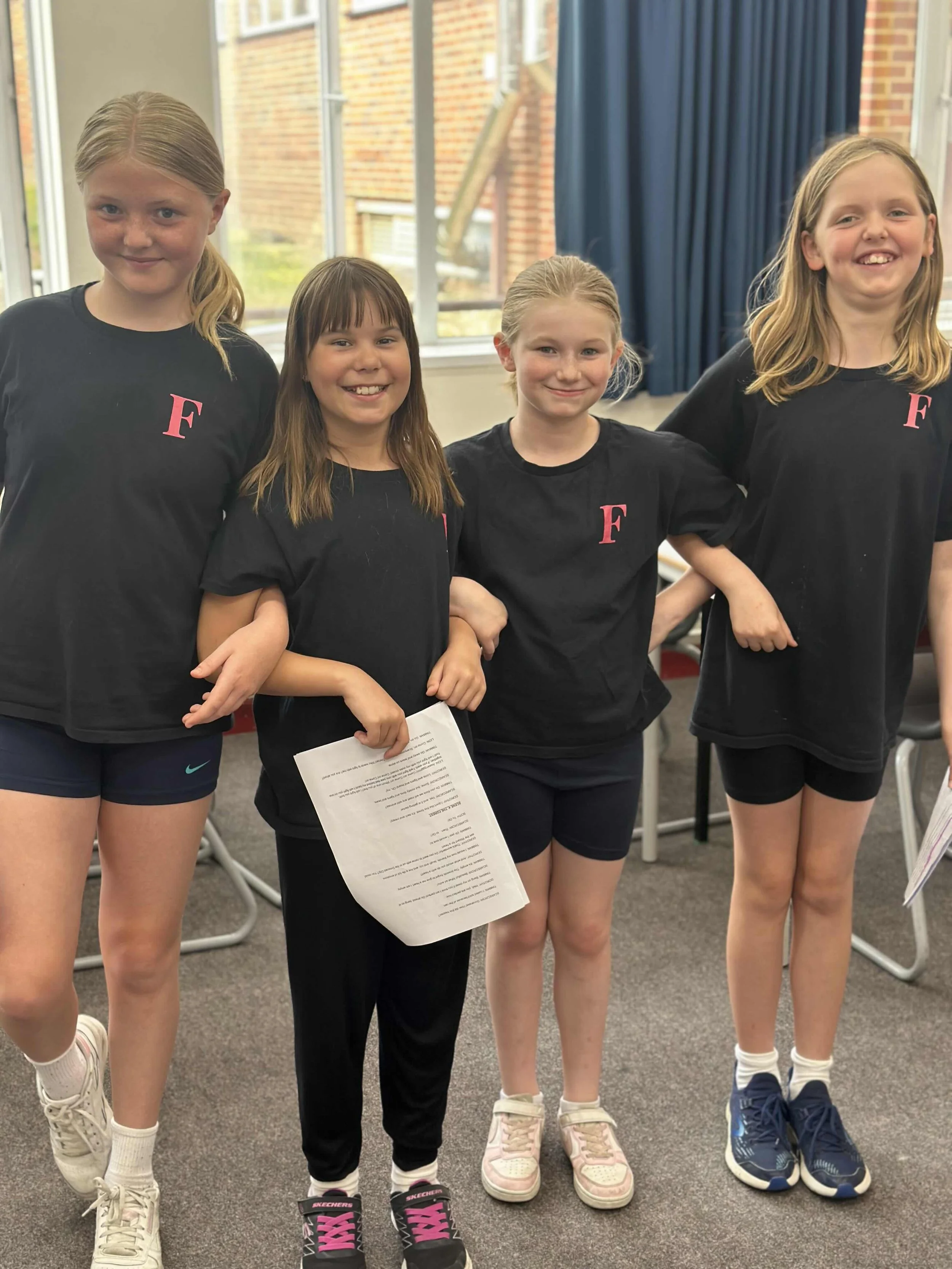 Four young girls standing together indoors, wearing matching black T-shirts with a pink letter 'F' and sporting shorts and sneakers, smiling at the camera.