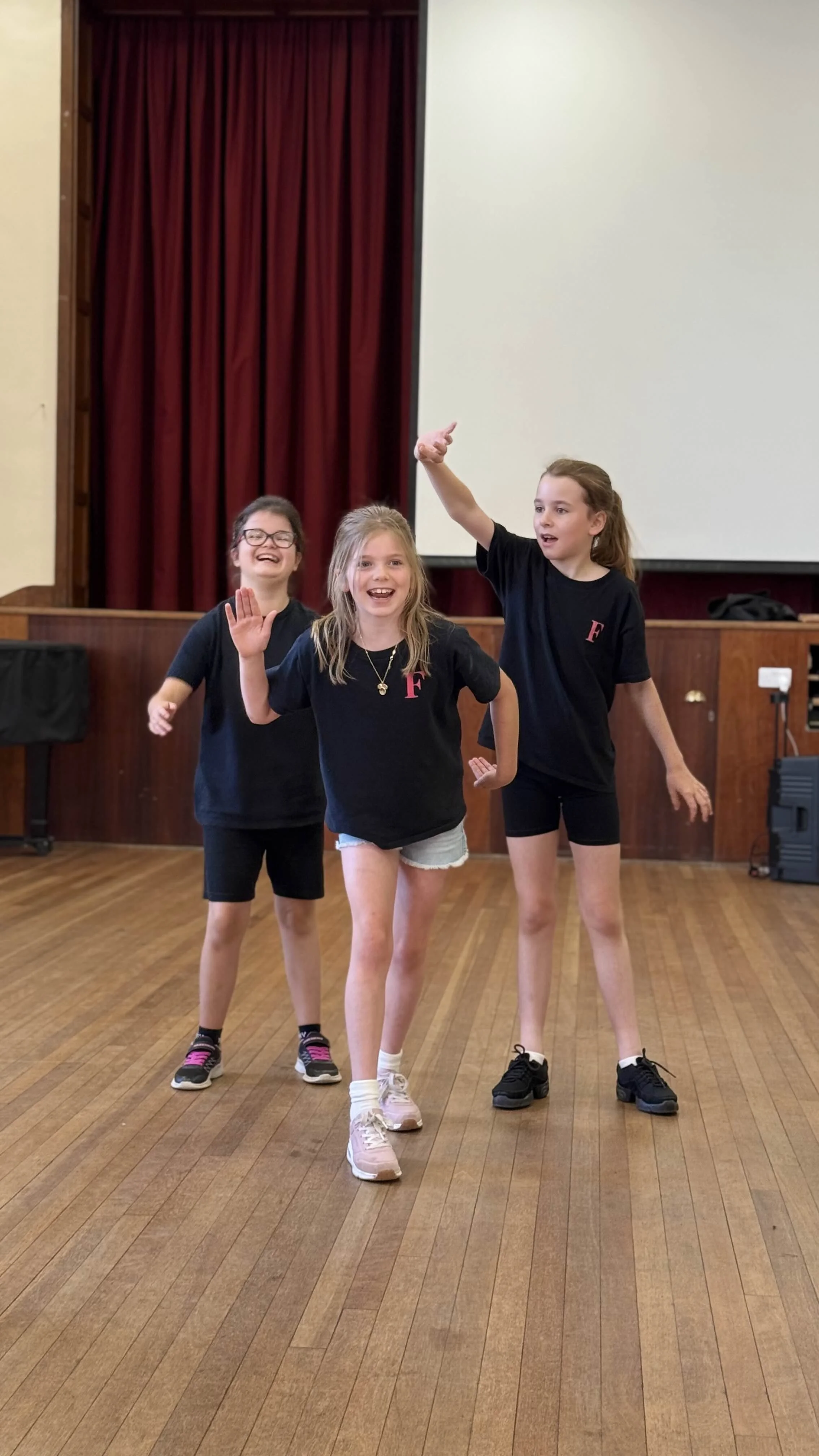 Three young girls dancing or performing on a wooden floor in a room with a red curtain and projector screen in the background.
