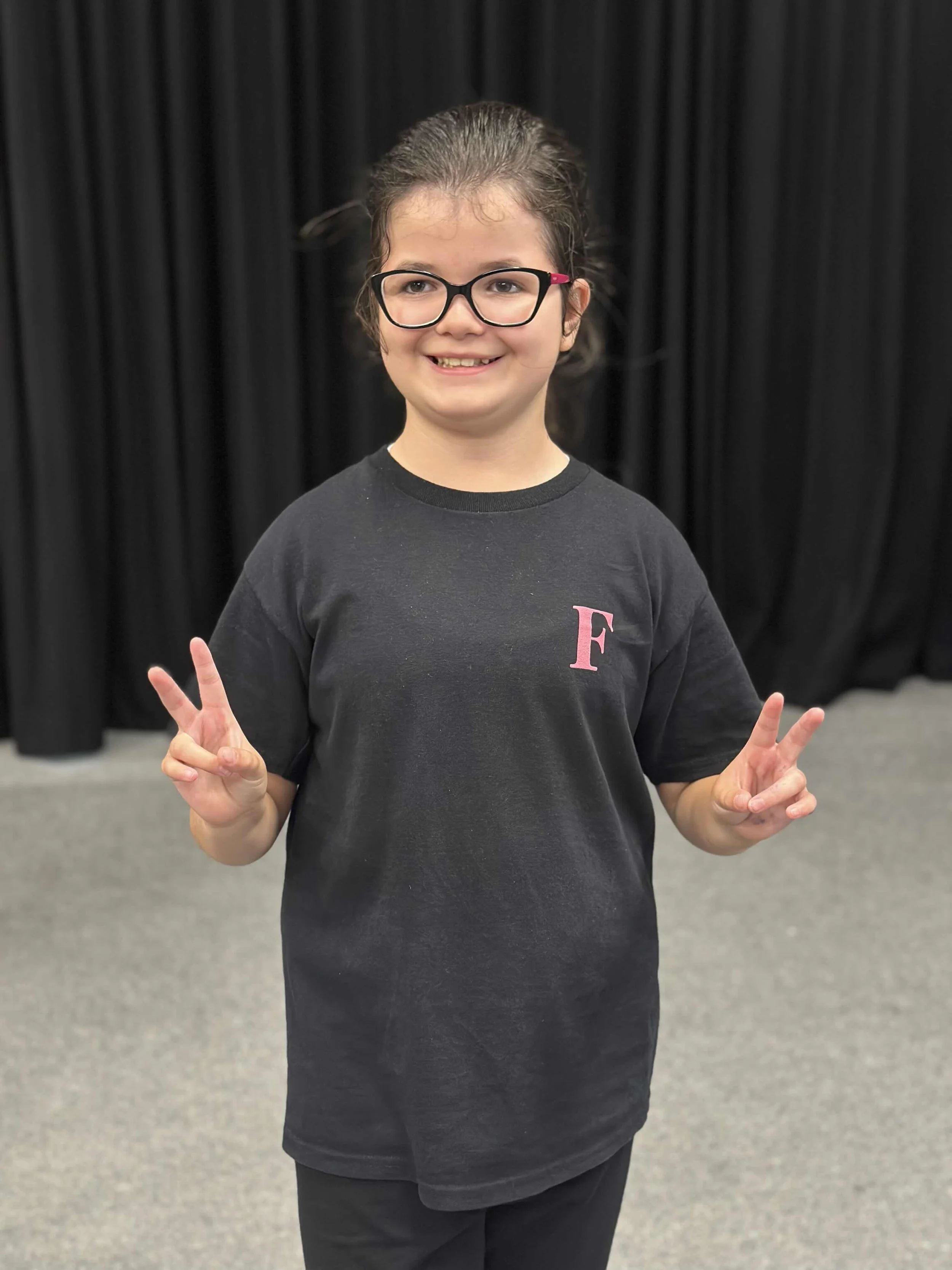 A young girl with glasses, wearing a black T-shirt with a pink letter 'F' on it, standing in front of a black curtain, smiling, and making peace signs with both hands.