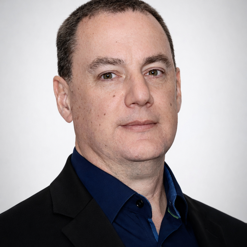 A man with short brown hair, wearing a dark suit jacket and a navy shirt, posing in front of a plain light background.