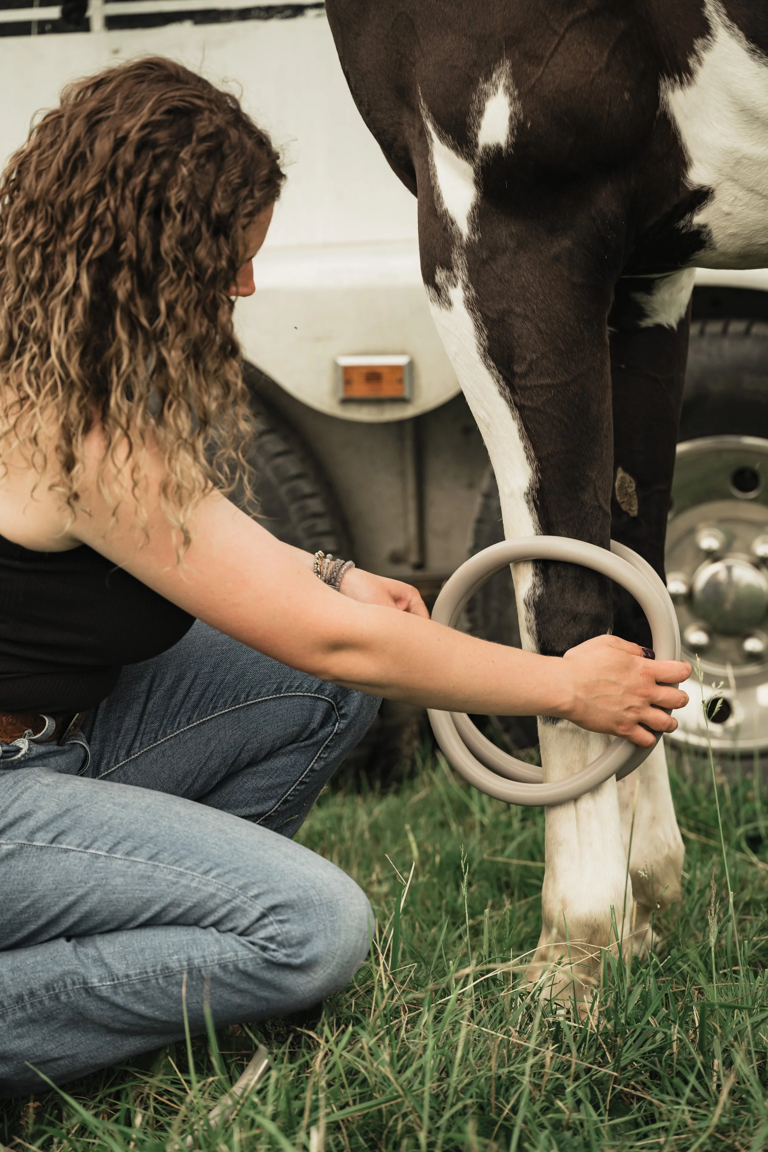 A woman kneeling on grass, fastening a large circle around the leg of a black and white horse, with a white vehicle in the background.