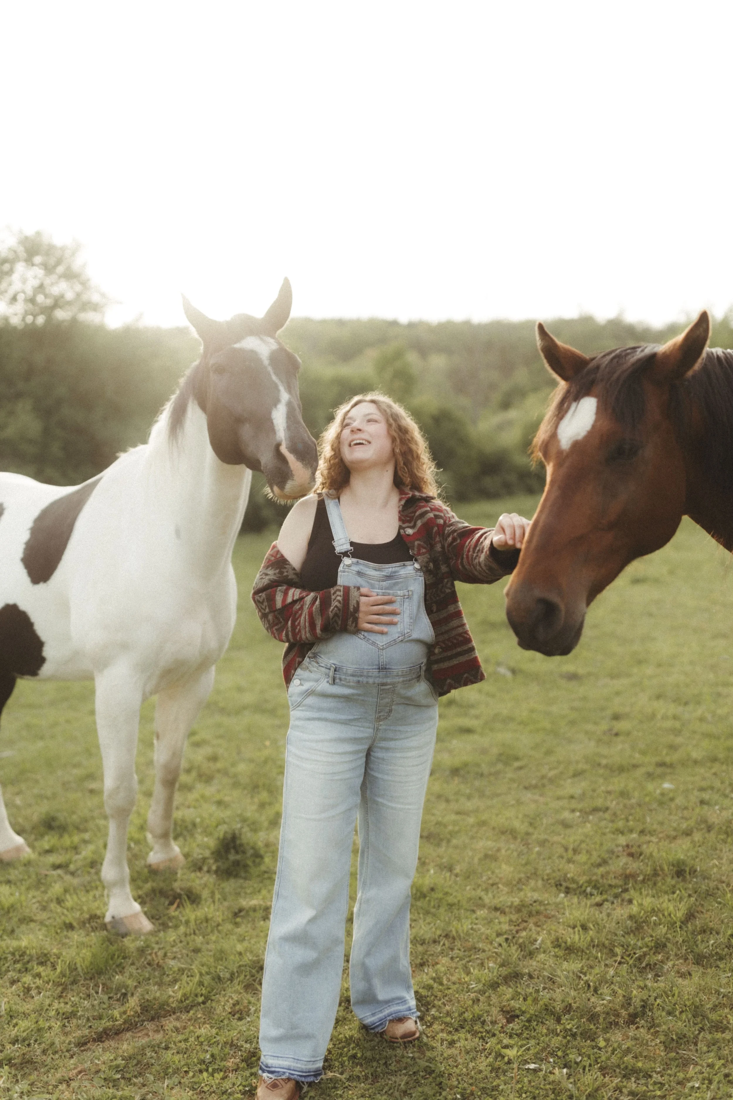 A woman with curly hair dressed in overalls and a jacket is interacting with two horses in a grassy field during sunset, smiling and enjoying the moment.
