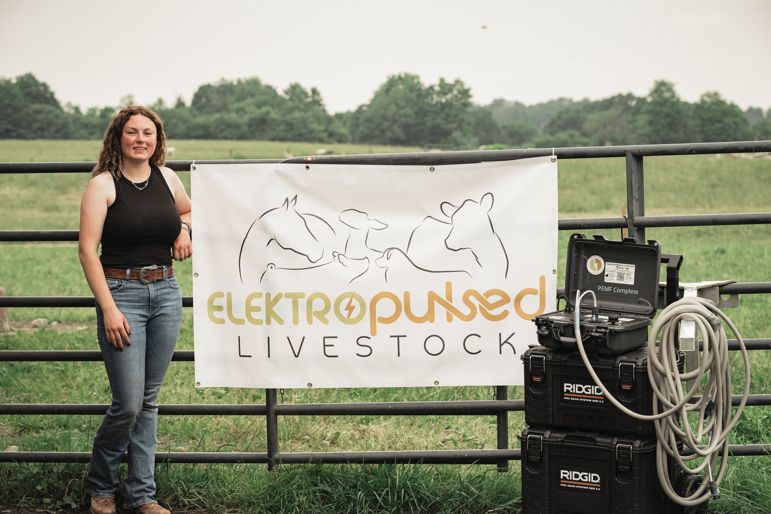 A woman standing by a fence with a sign that reads 'ELEKROPULSED LIVESTOCK' and an illustration of farm animals. There is equipment with cables on the ground.