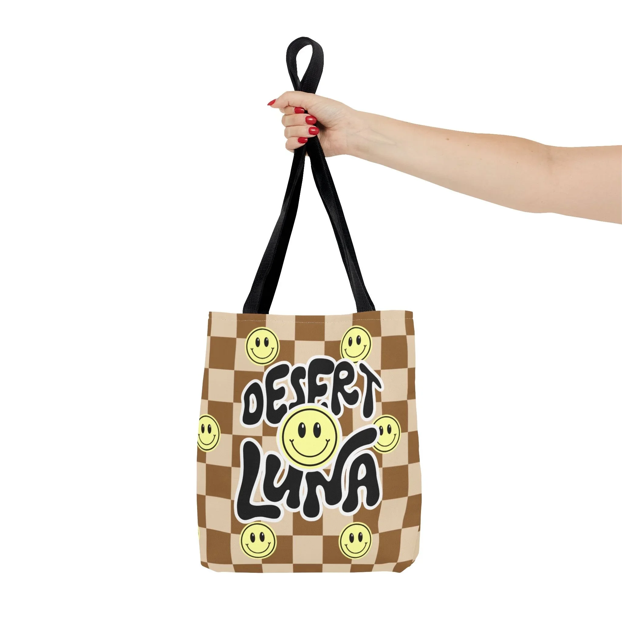 Hand holding a brown and beige checkered tote bag with smiley face designs and the words "Desert Luna" on the front