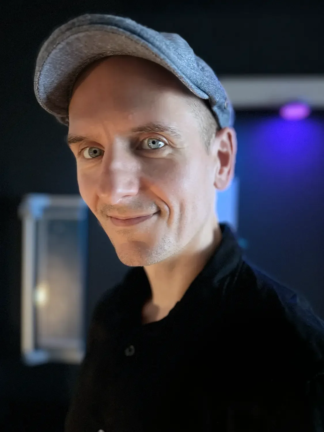 Close-up of a man wearing a gray baseball cap, smiling, with a dark background and purple lighting