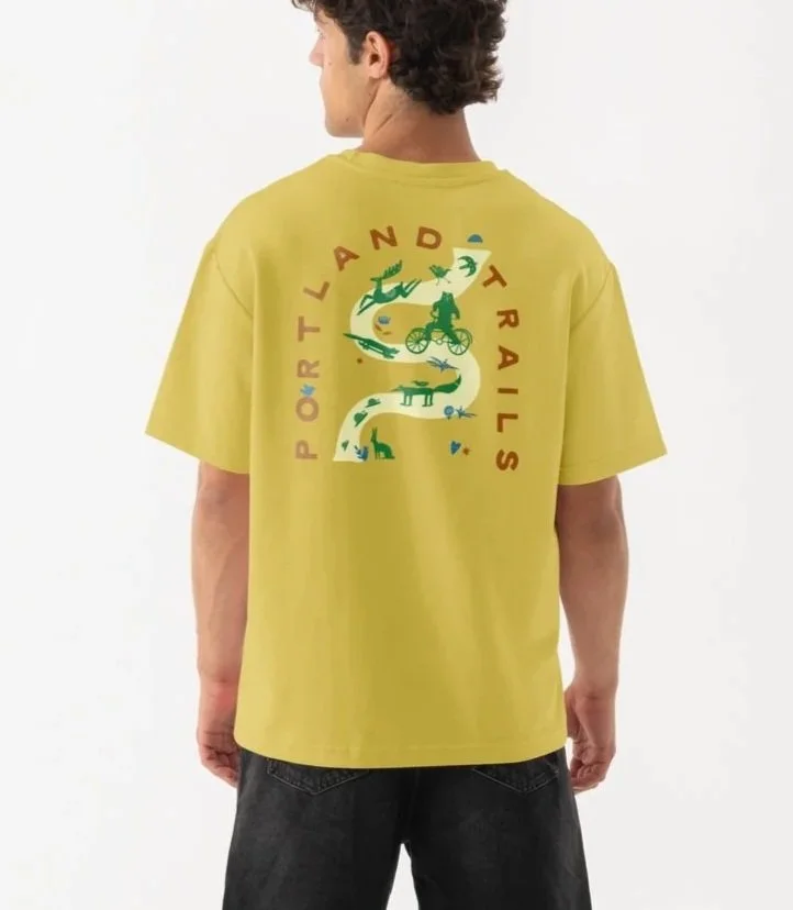 Back view of a young man standing on white background, wearing a yellow T-shirt with a colorful graphic and the words 'PORTLAND TRAILS' on the back, black shorts, and white sneakers.