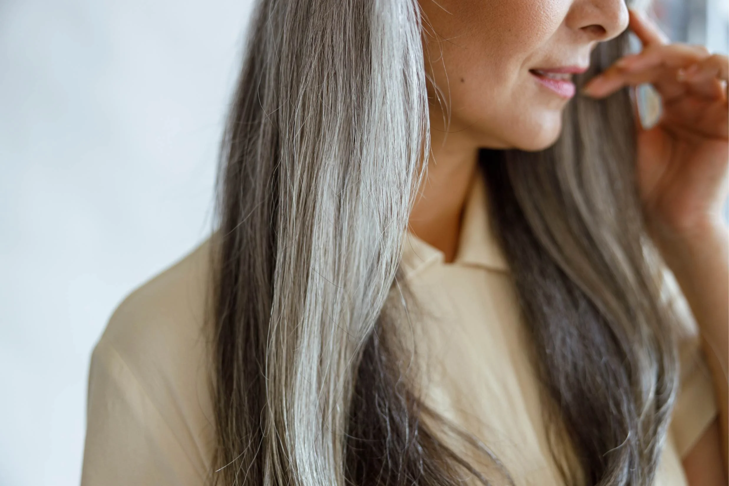 Close-up of a woman with long gray hair and light skin, smiling gently, wearing a beige shirt, indoors.