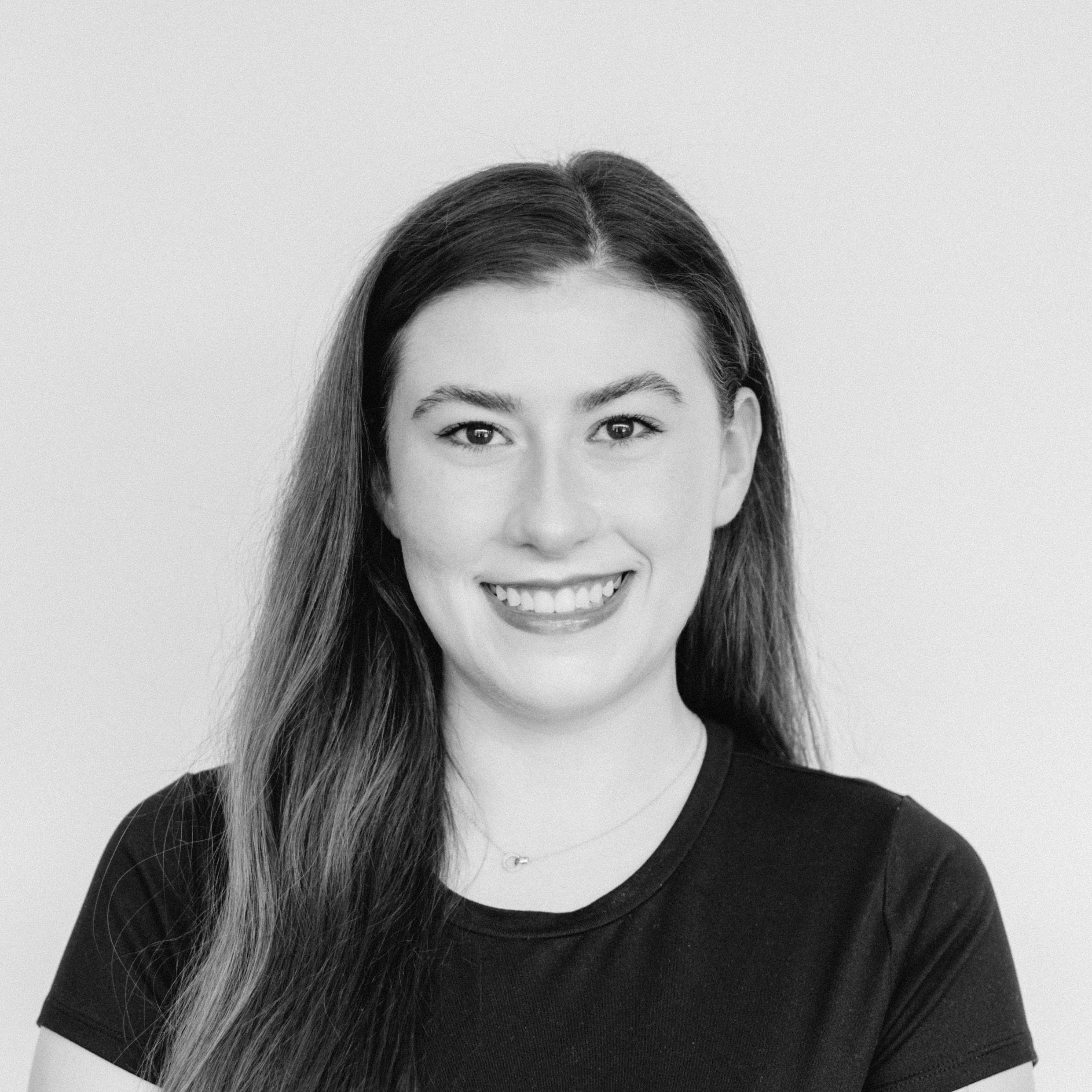 A smiling young woman with light skin tone and long dark hair wearing a black top, against a plain background.