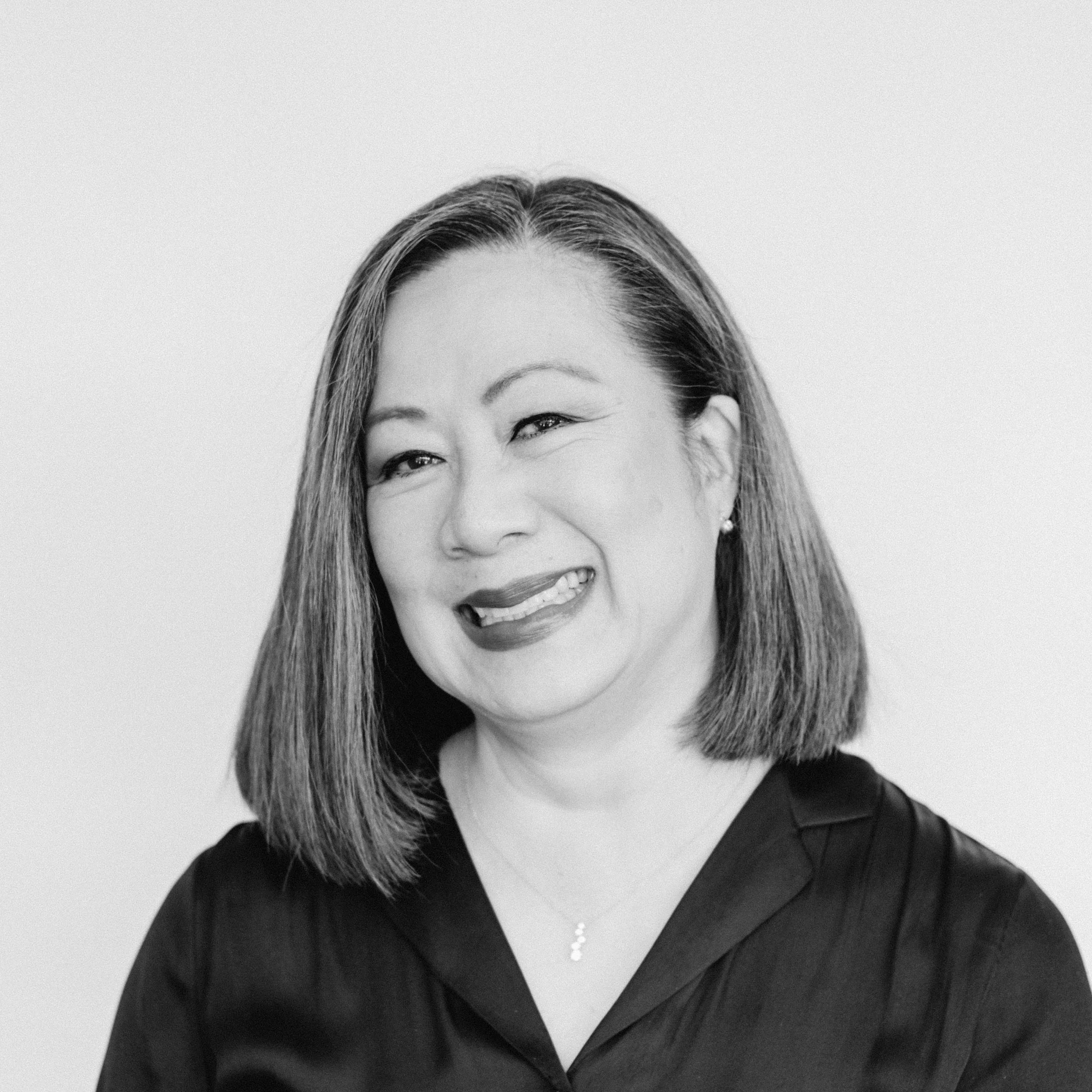 A smiling woman with medium-light skin tone and shoulder-length black hair, wearing a dark blouse and a necklace, against a plain background.
