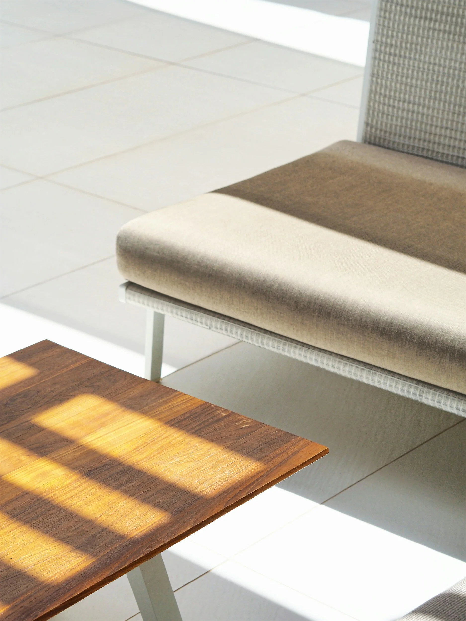 Close-up of a beige cushioned chair and a wooden coffee table with shadows cast by window blind slats on its surface, in a bright waiting-room setting.