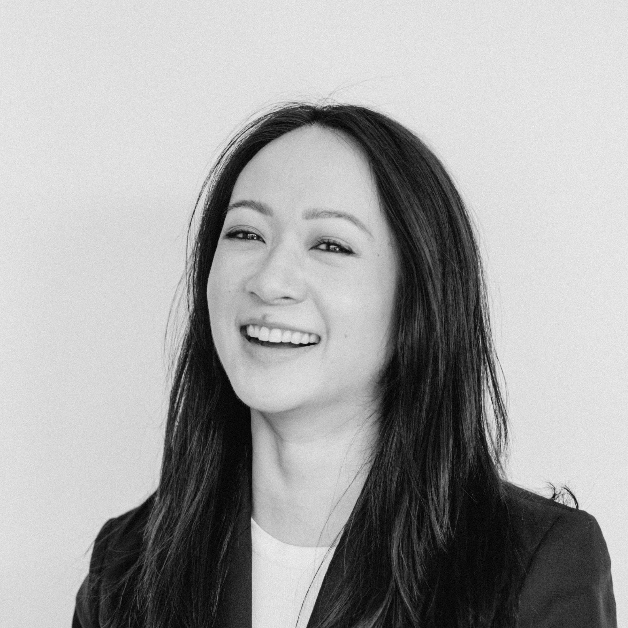 A laughing woman with medium-light skin tone and long dark hair, wearing a blazer and white top, against a plain light background.