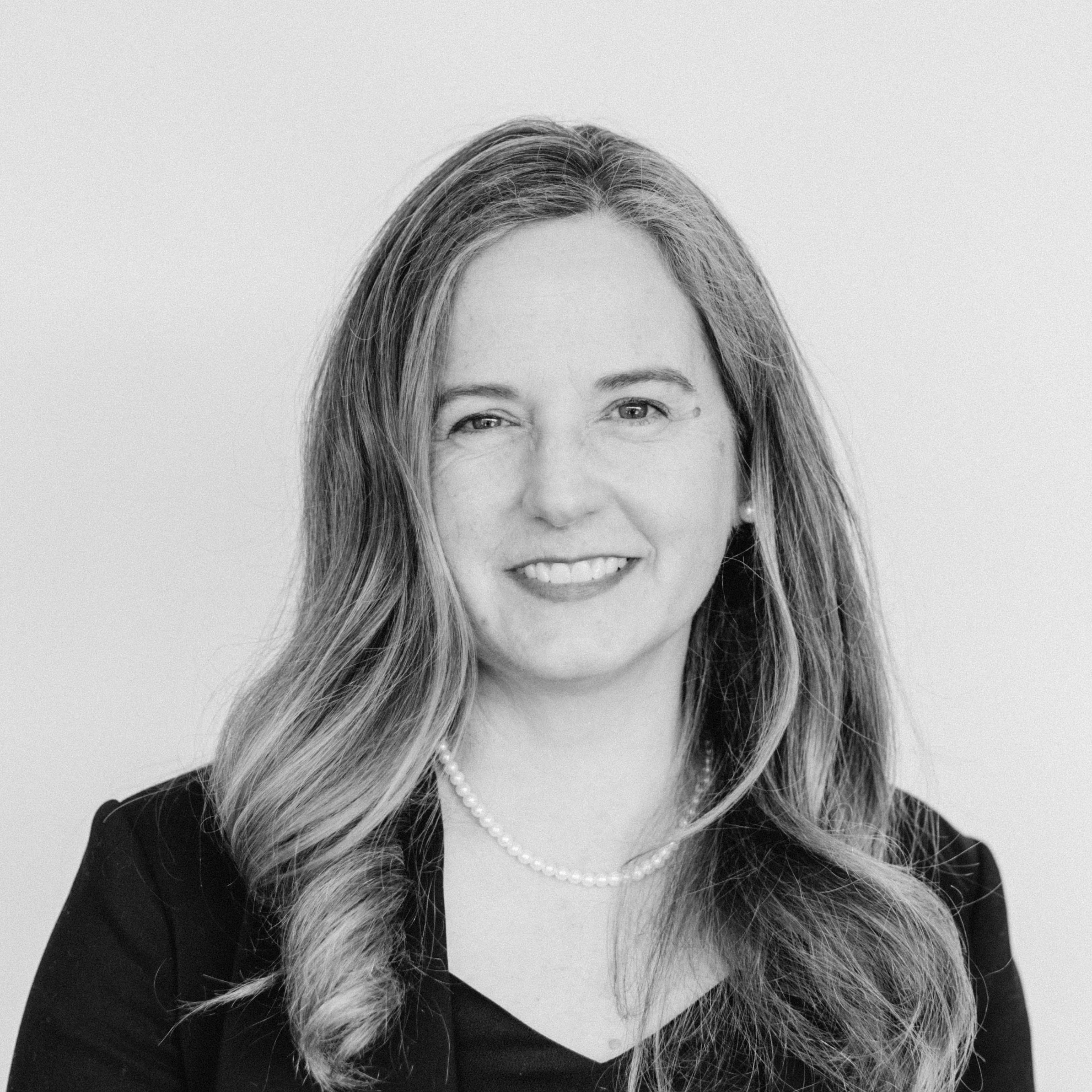 A smiling woman with light skin tone and light-brown wavy hair, wearing a pearl necklace and a dark blazer.