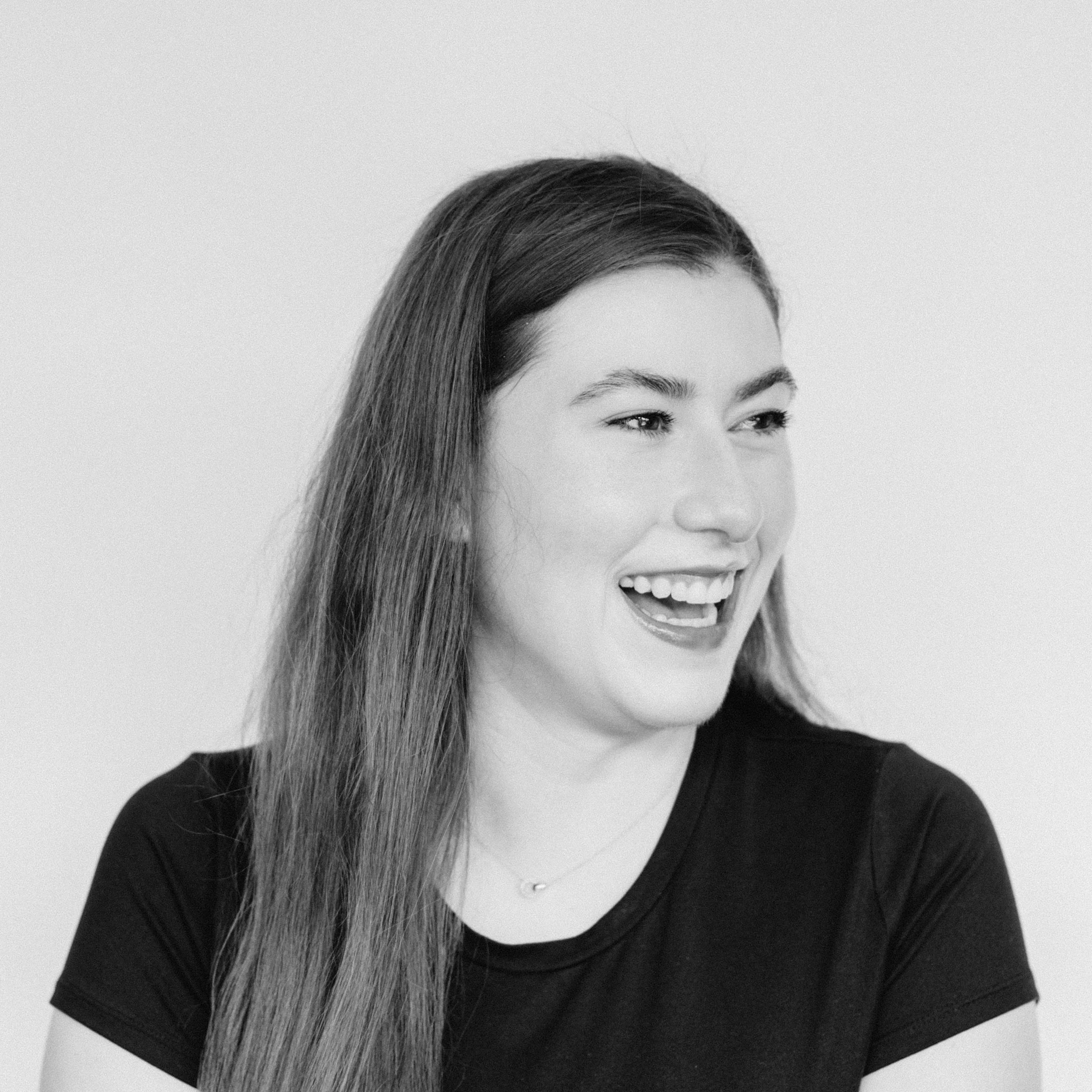 A smiling young woman with long hair, laughing and looking to her right, wearing a black top, against a plain light background.