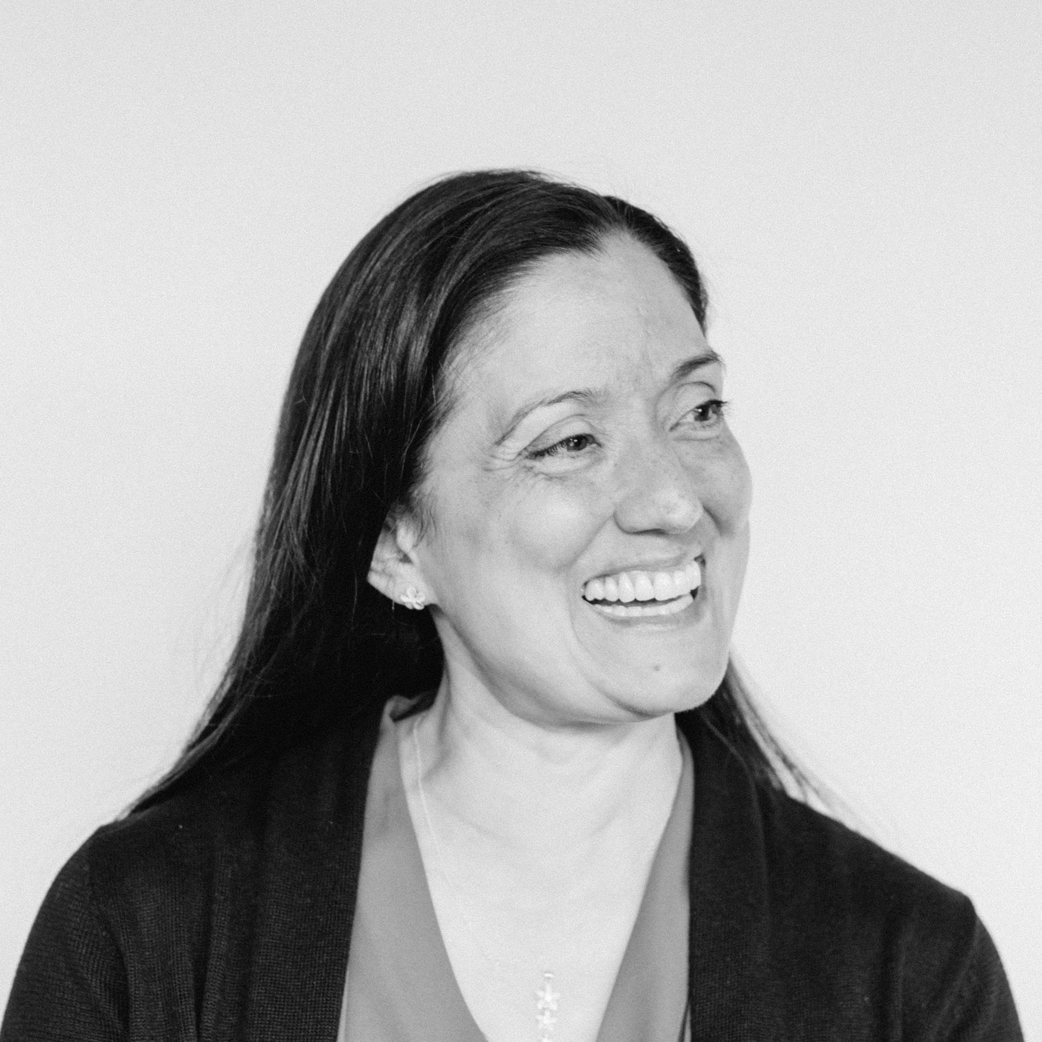 A laughing woman with medium-light skin tone and long dark hair, wearing a dark cardigan and a necklace, against a plain background.