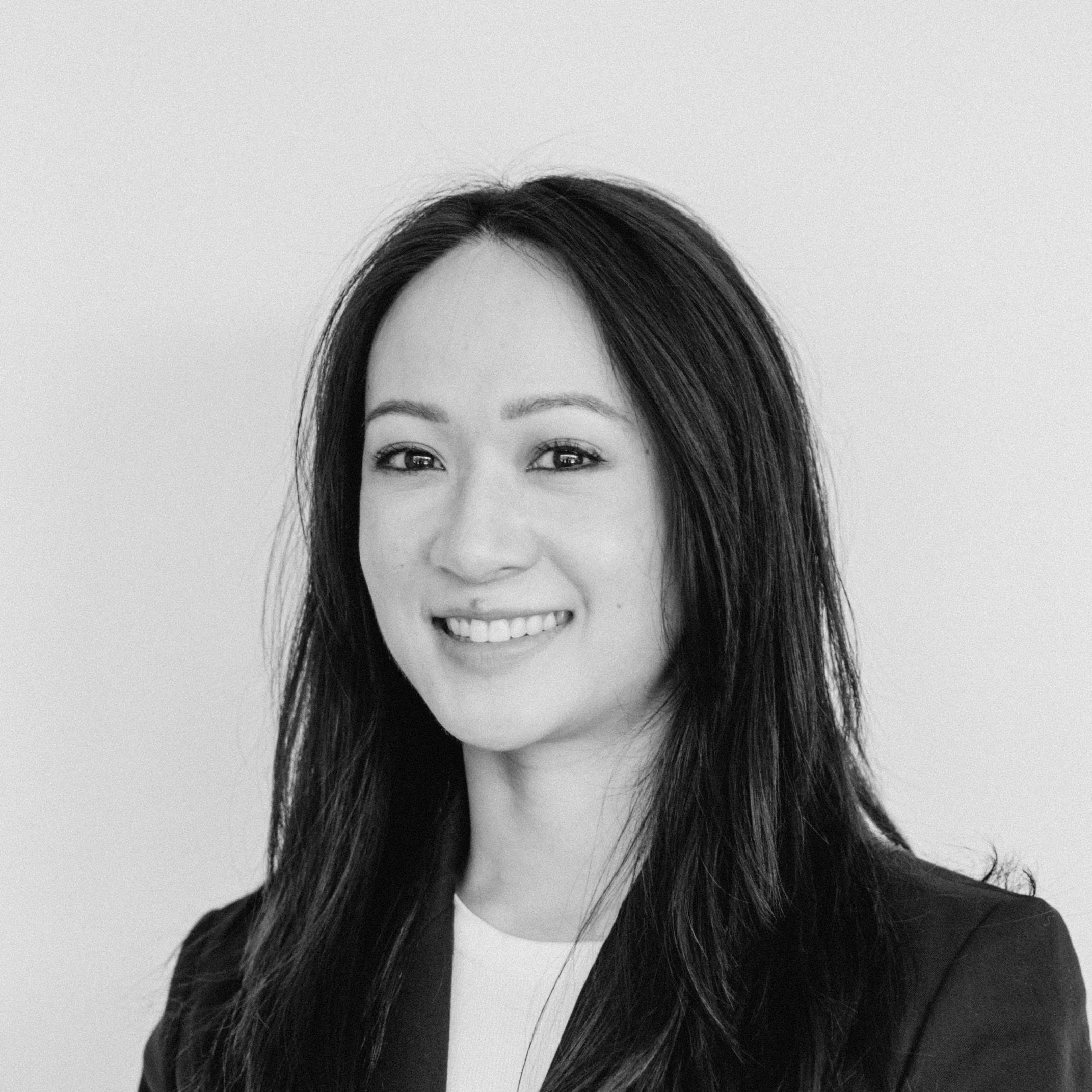 A smiling woman with medium-light skin tone and long dark hair wearing a blazer and white top against a light background.