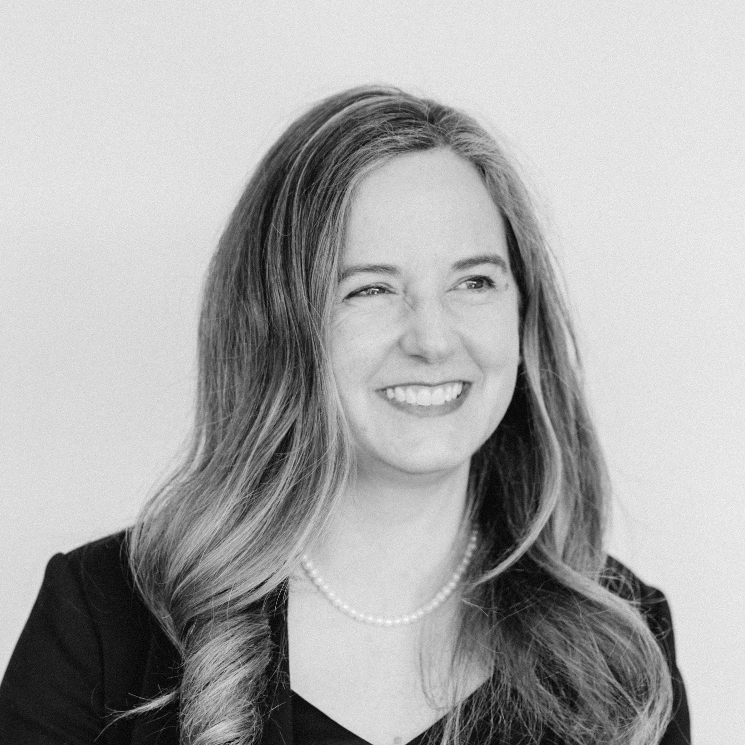 A smiling woman with light skin tone and light-brown wavy hair, wearing a pearl necklace and a dark blazer.