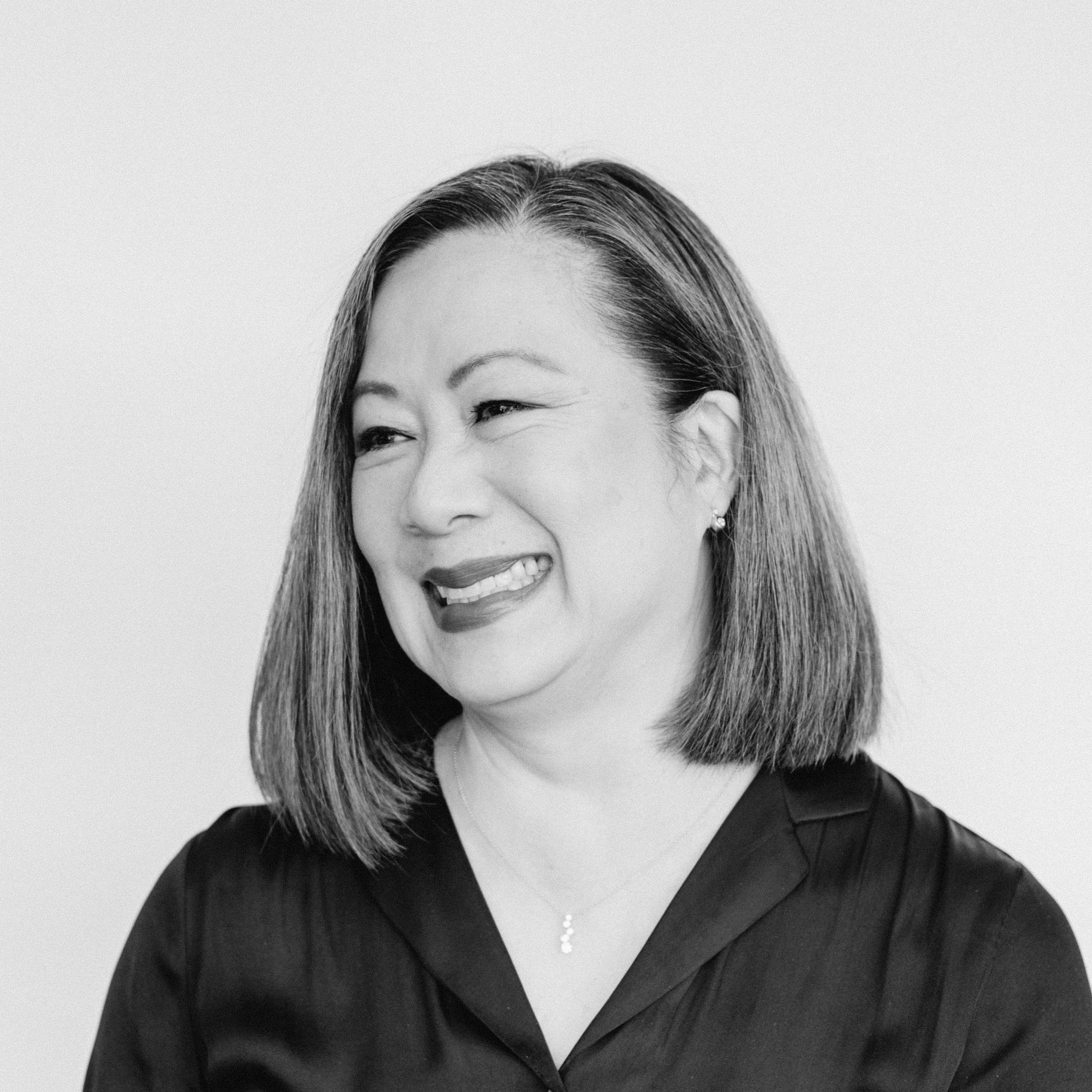 A smiling woman with medium-light skin tone and shoulder-length black hair, wearing a dark blouse and a necklace, against a plain background.