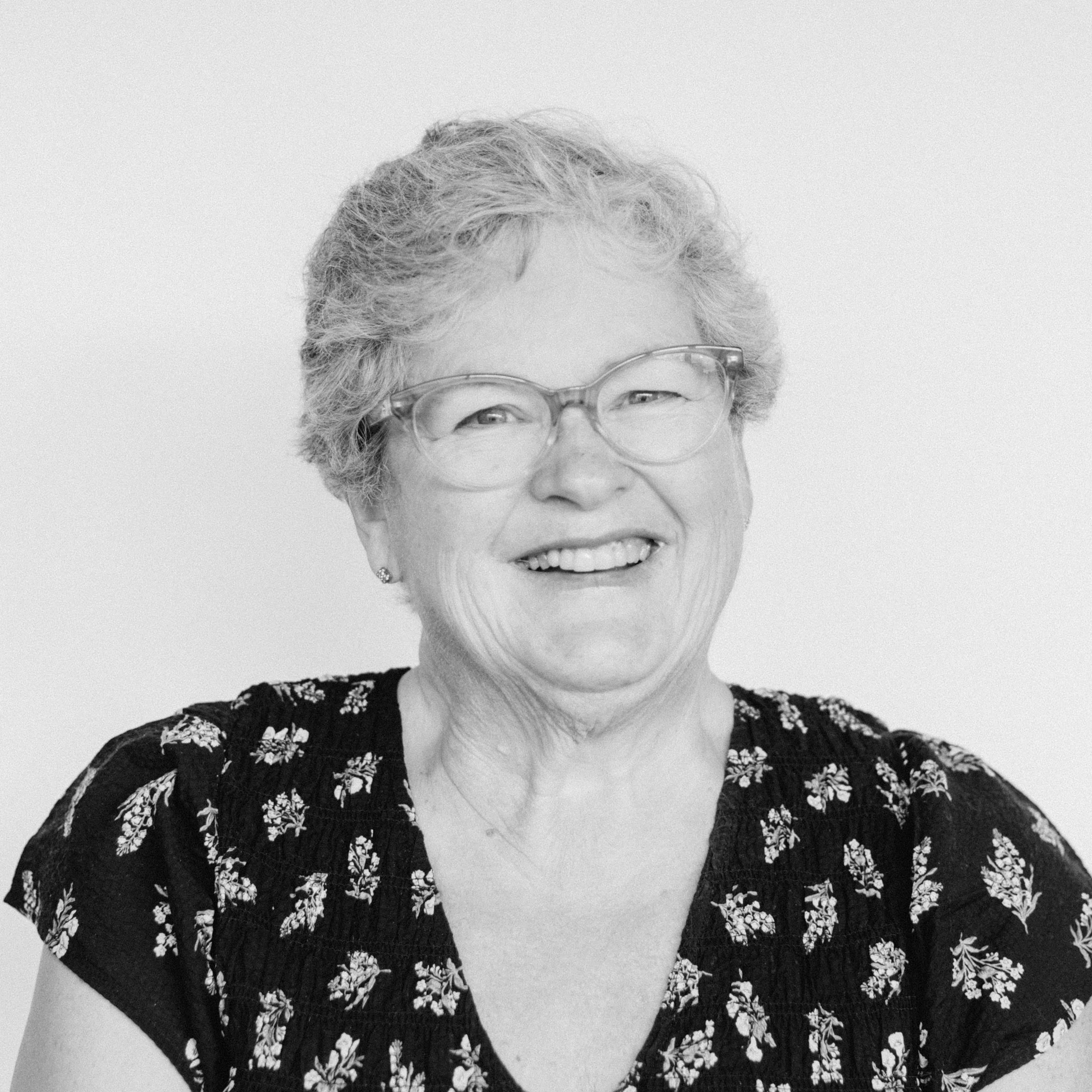 A smiling older woman with light skin tone and short curly gray hair, wearing glasses and a floral blouse.
