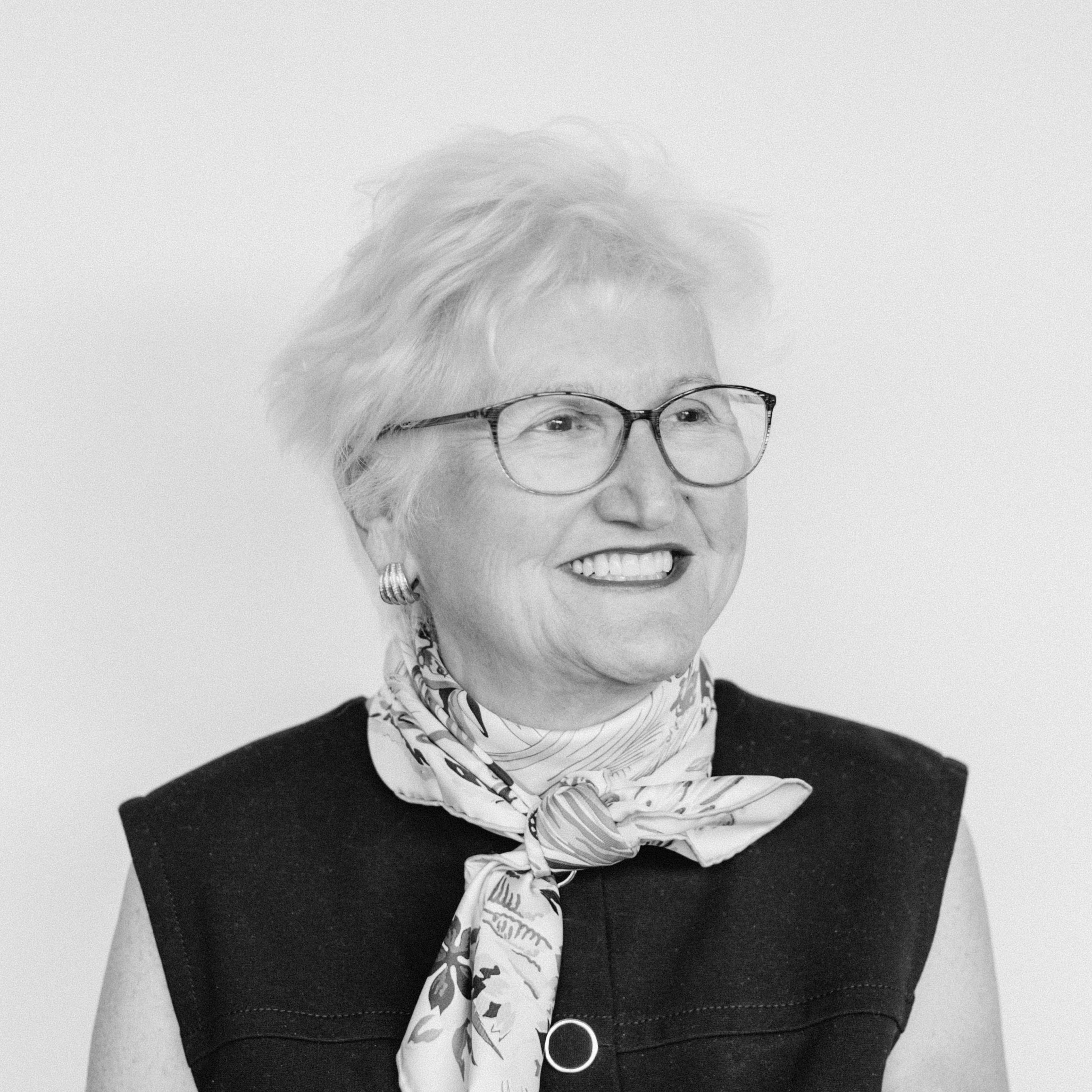 An older smiling woman with light skin tone and short white hair wearing glasses, earrings, a patterned scarf, and a sleeveless top.