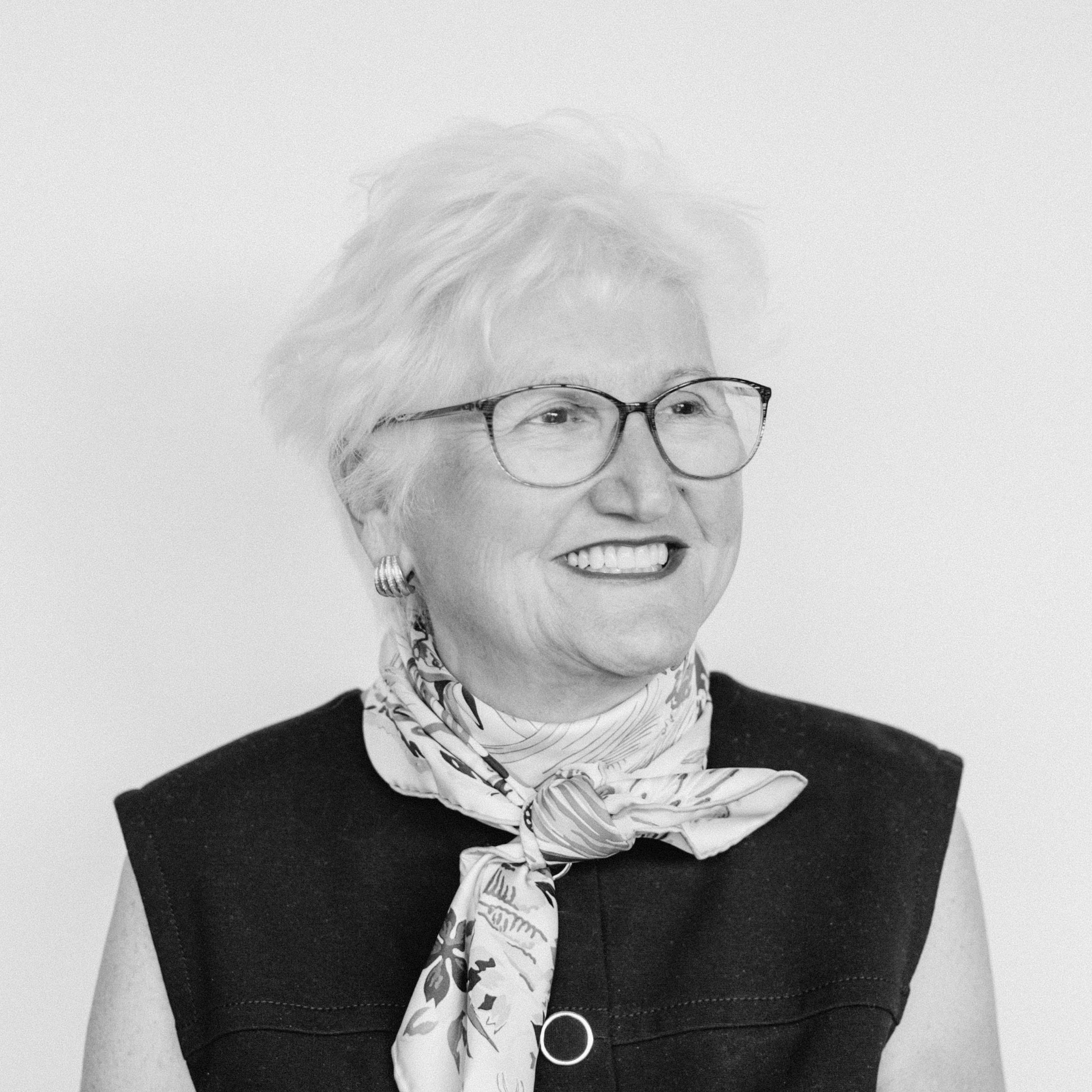 An older smiling woman with light skin tone and short white hair wearing glasses, earrings, a patterned scarf, and a sleeveless top.
