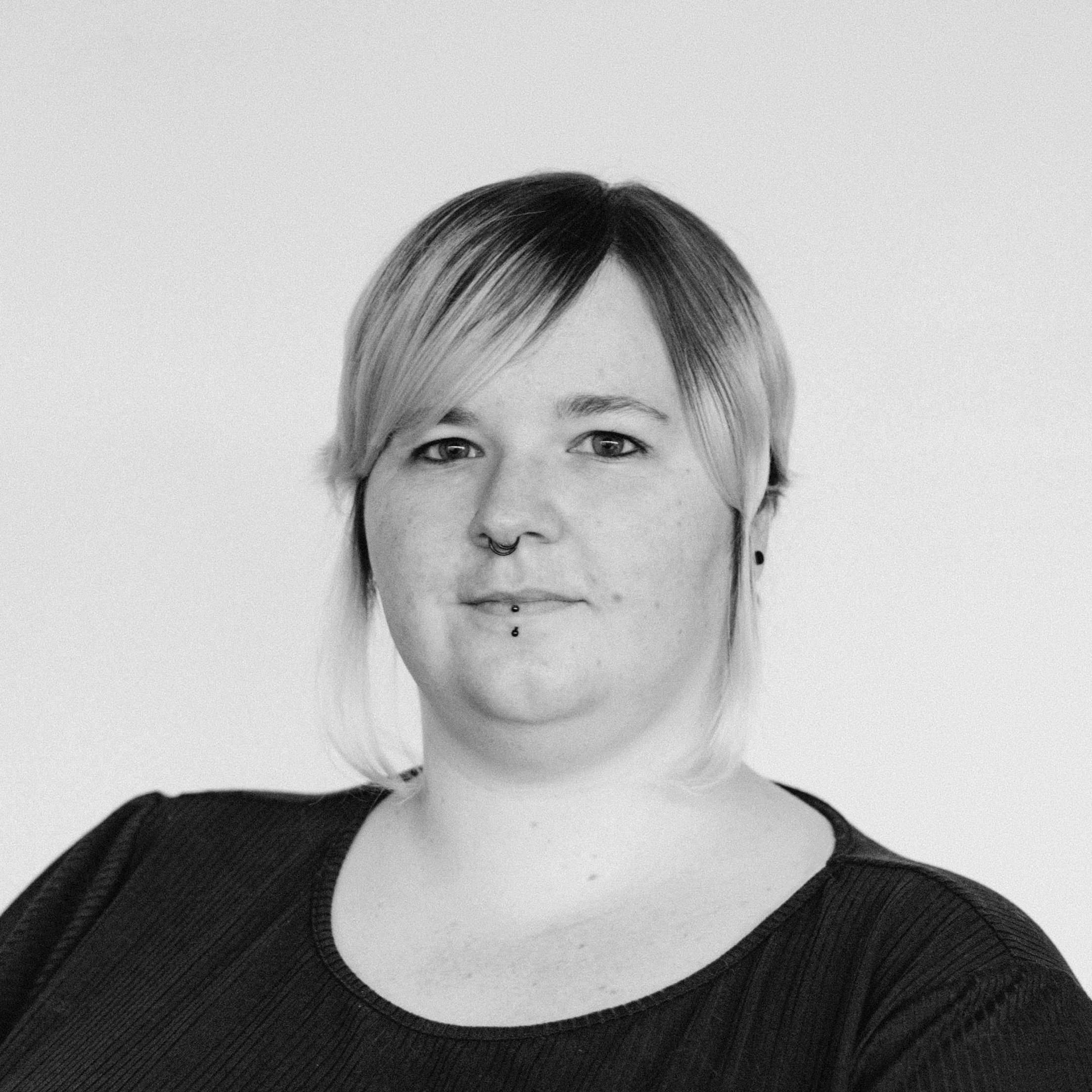A woman with light skin tone, short light hair, side-swept bangs, a nose ring, and a lip piercing, wearing a dark t-shirt.