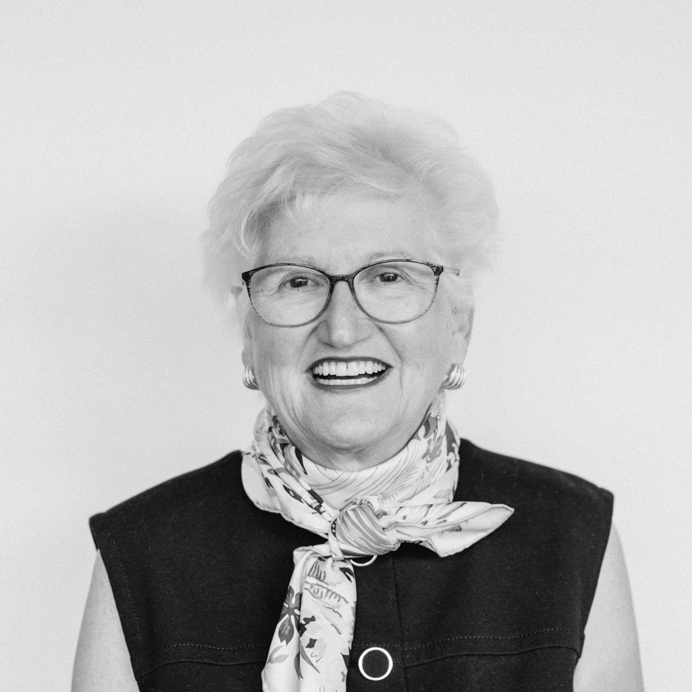 An older smiling woman with light skin tone and short white hair wearing glasses, earrings, a patterned scarf, and a sleeveless top.