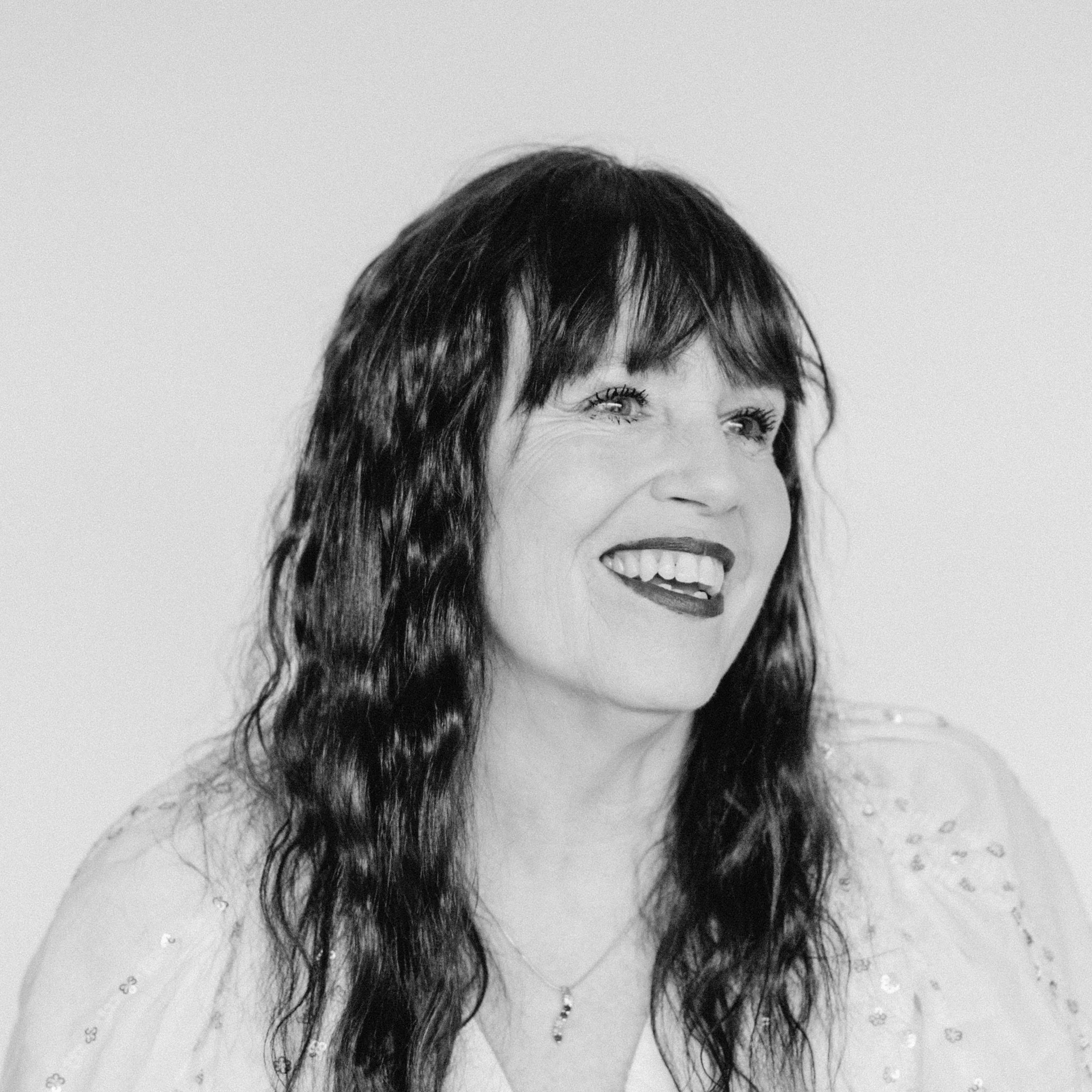 A smiling woman with light skin tone, curled bangs and wavy dark hair, wearing a light blouse and necklace.
