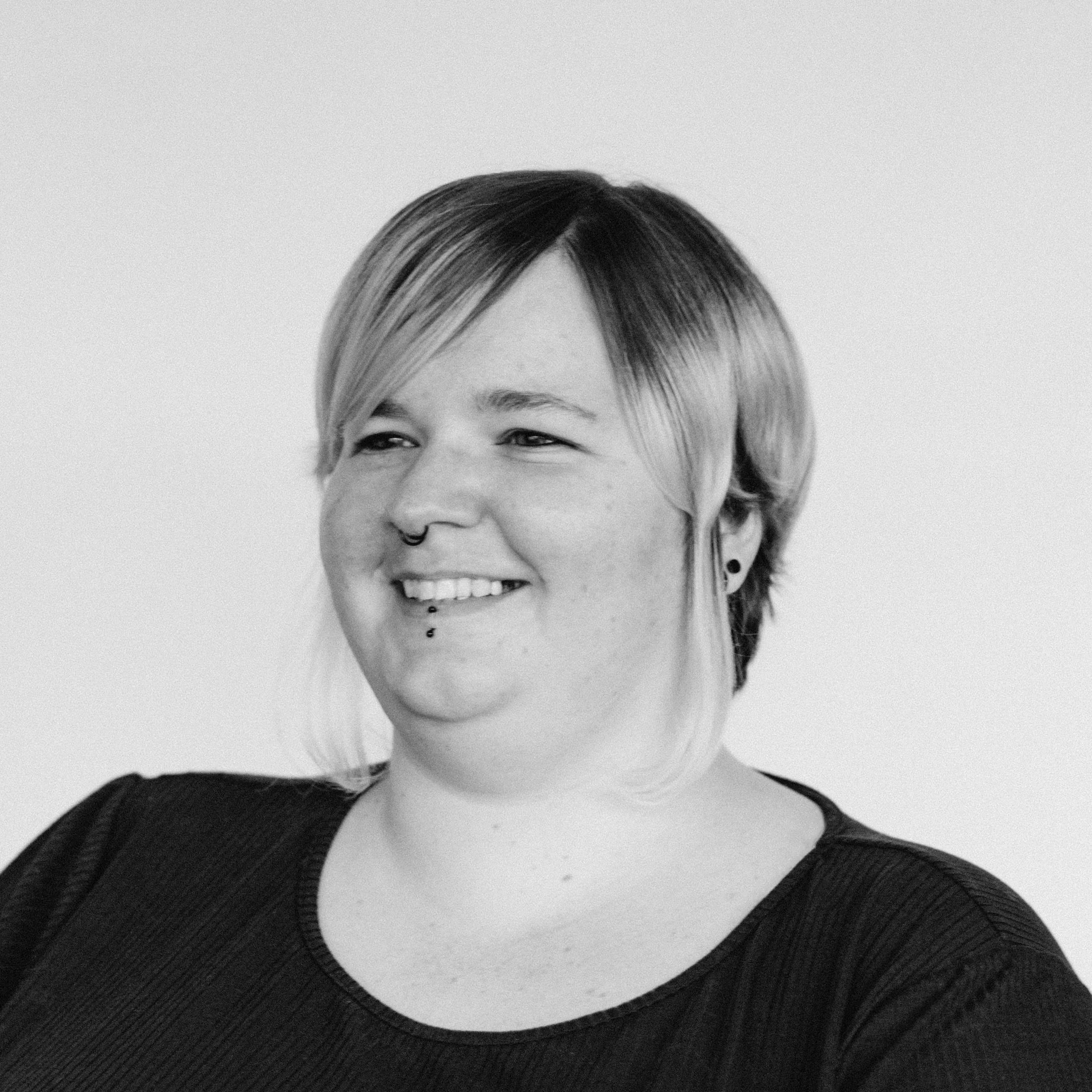 A smiling woman with light skin tone, short light hair, side-swept bangs, a nose ring, and a lip piercing, wearing a dark t-shirt.