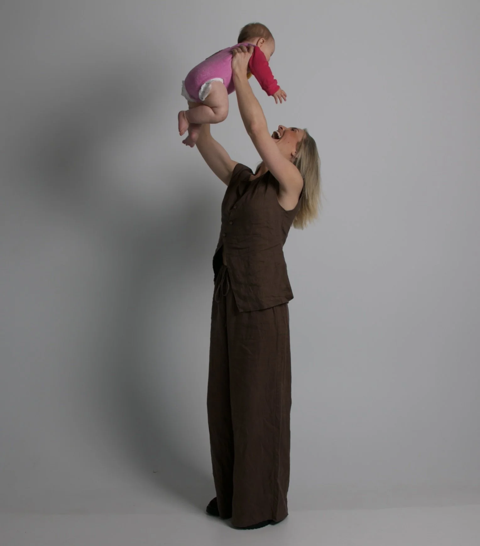 Woman holding a smiling baby up in the air against a plain gray background.