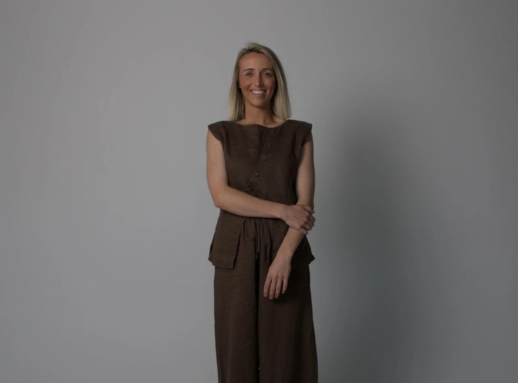 A woman with shoulder-length blonde hair, smiling and standing with arms crossed, wearing a sleeveless brown top and matching wide-leg pants, against a plain gray background.