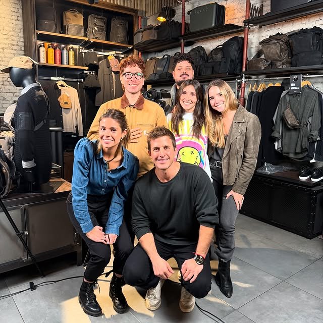Seis personas posando y sonriendo en una tienda de ropa con estantes llenos de bolsas y prendas oscuras.