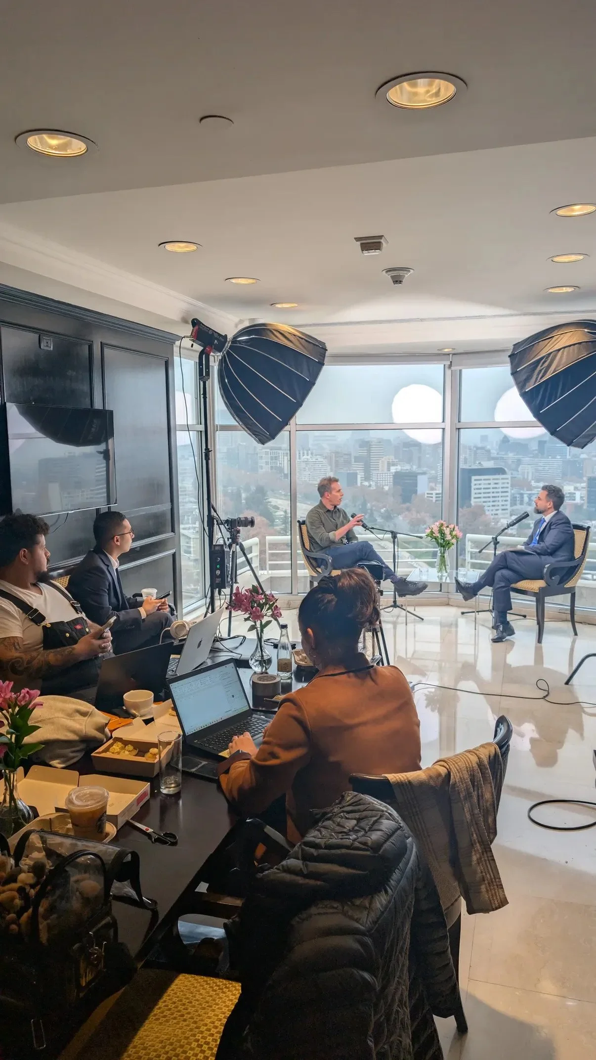 Escena de una entrevista en un estudio con vistas a la ciudad, con varias personas, cámaras, iluminación profesional y laptops en una mesa.