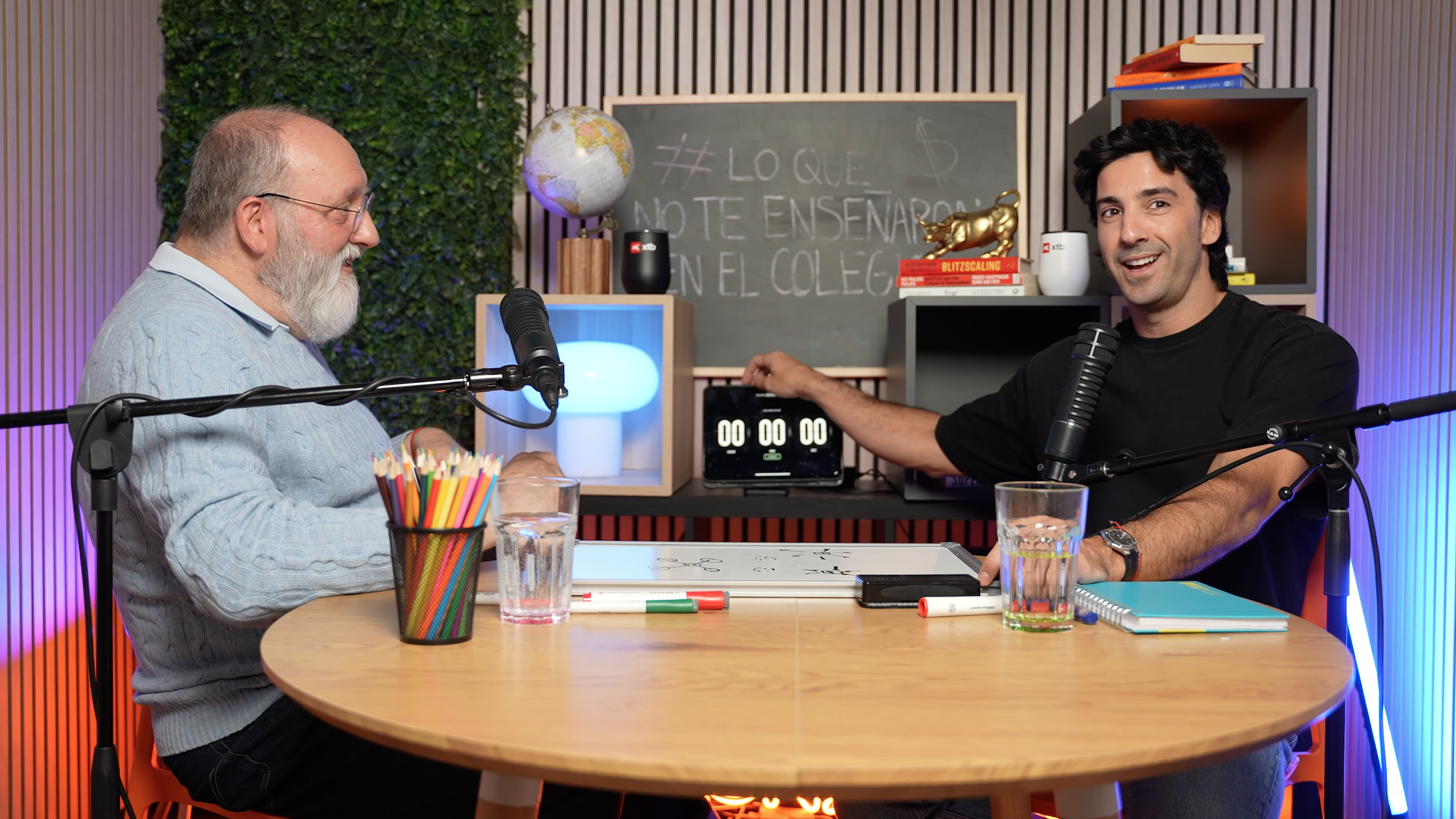 Dos hombres en un estudio grabando un podcast, con una mesa, micrófonos, y libros, uno de ellos tocas un interruptor en una caja con pantalla digital, y en el fondo un pizarrón con escritura en español.
