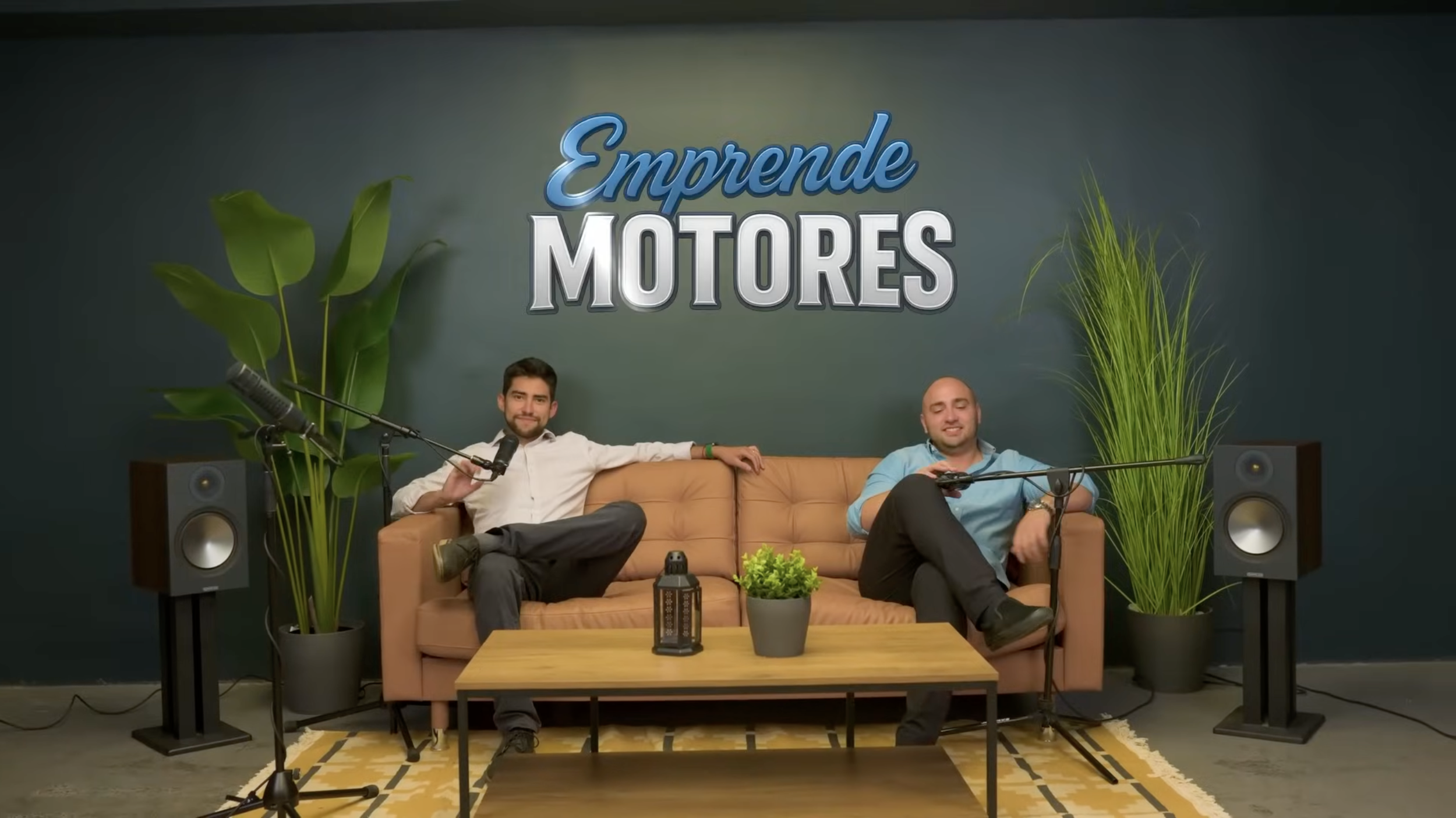 Dos hombres sentados en un sillón de color marrón, rodeados de plantas y equipo de grabación, en un estudio con un cartel que dice "Emprende Motores" en la pared