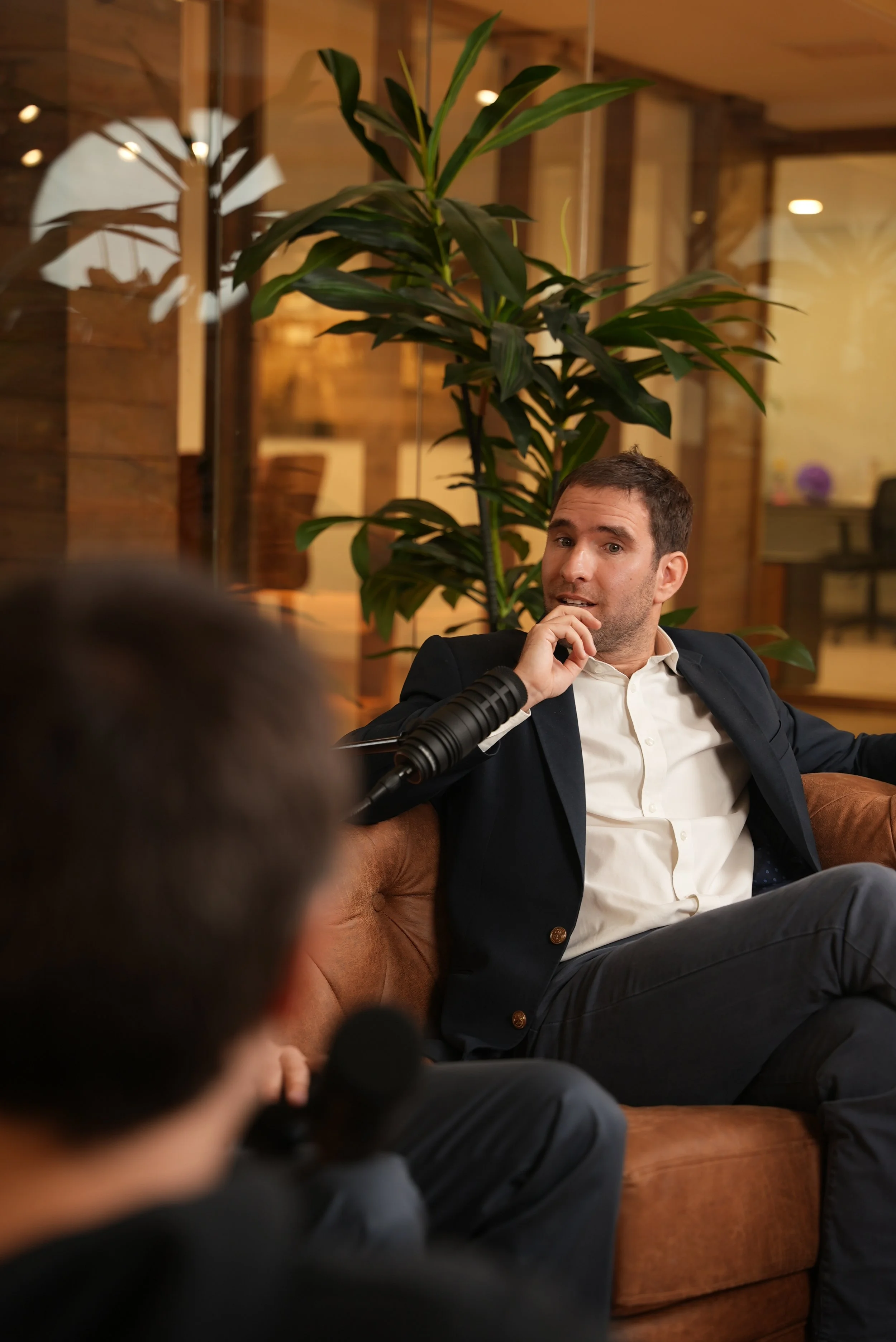 Dos hombres en una conversación en un ambiente moderno, uno de ellos está sentado en un sofá de color marrón y el otro en primer plano sosteniendo un micrófono, con plantas y paredes de madera en el fondo.