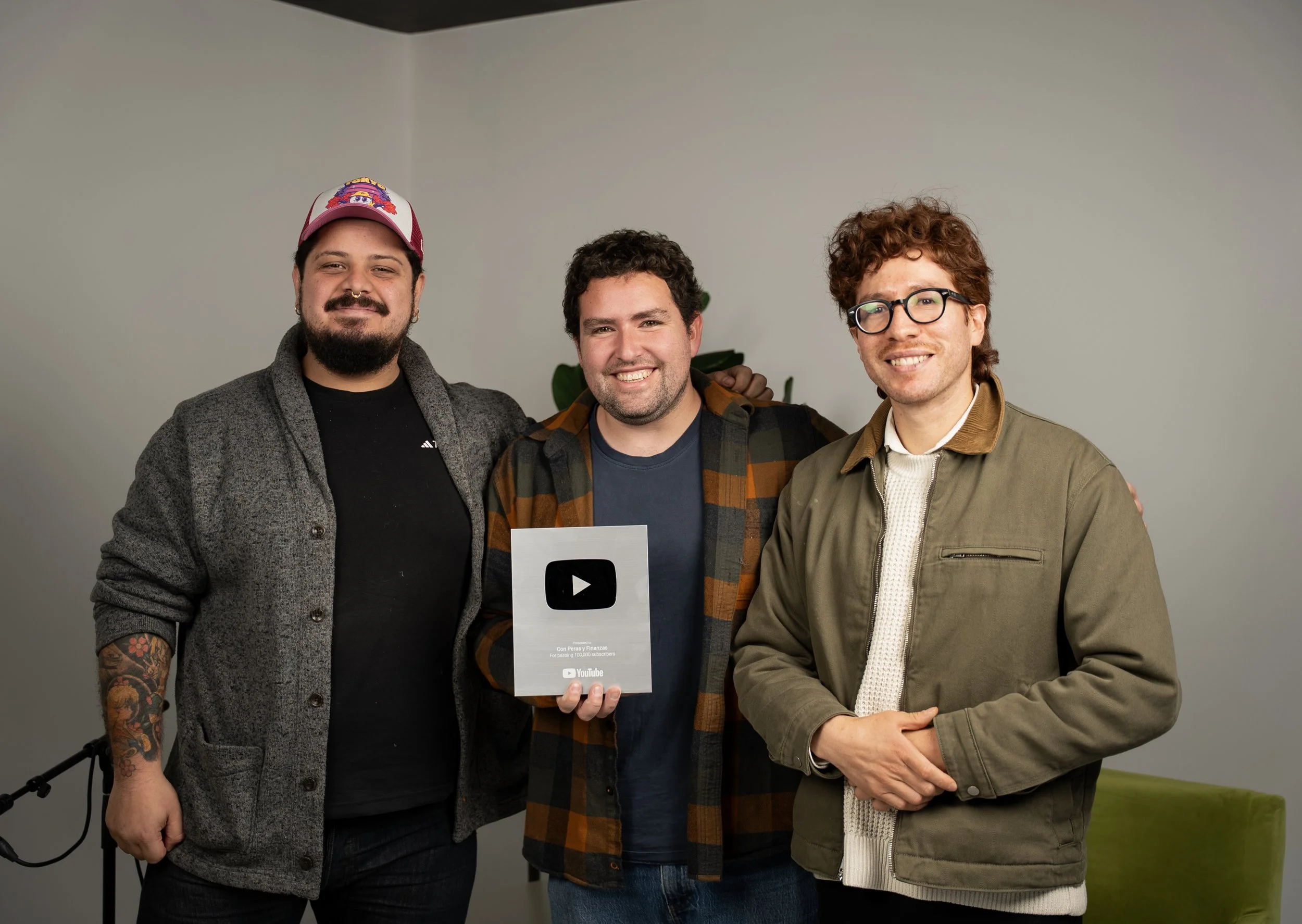 Tres hombres sonriendo y posando con un premio de YouTube, en un fondo simple y una planta.