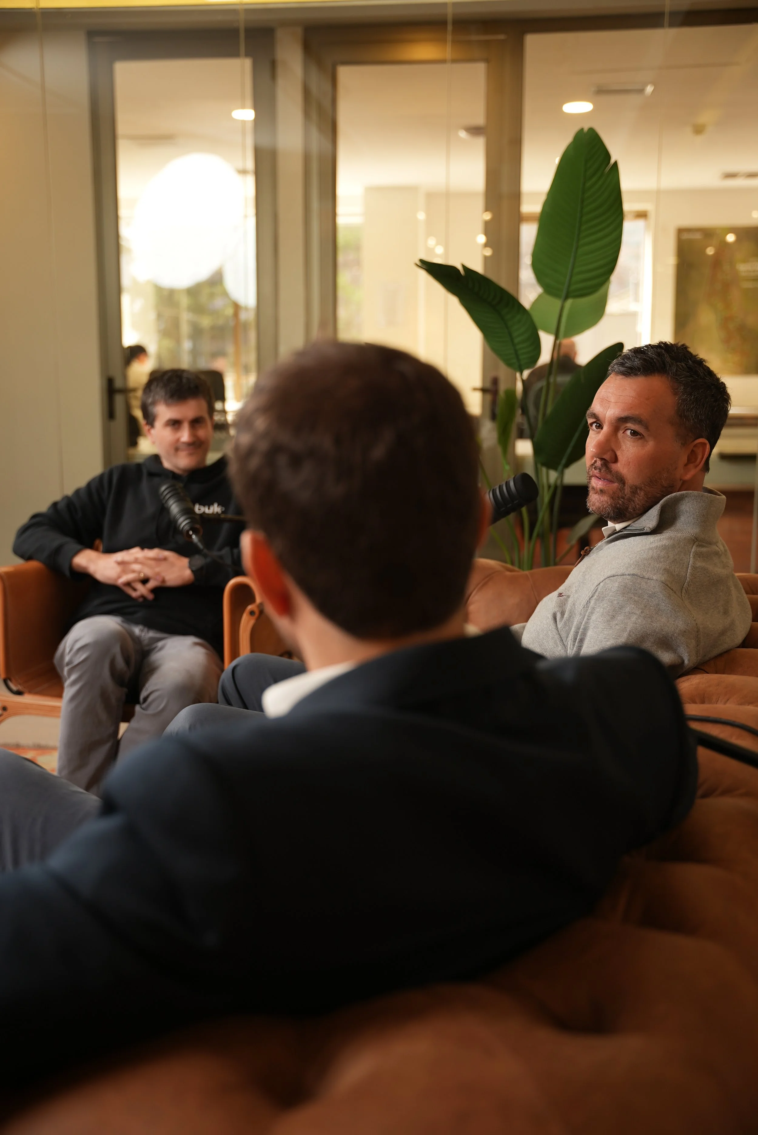 Tres hombres en una reunión o entrevista en un ambiente interior con plantas y ventanas grandes.