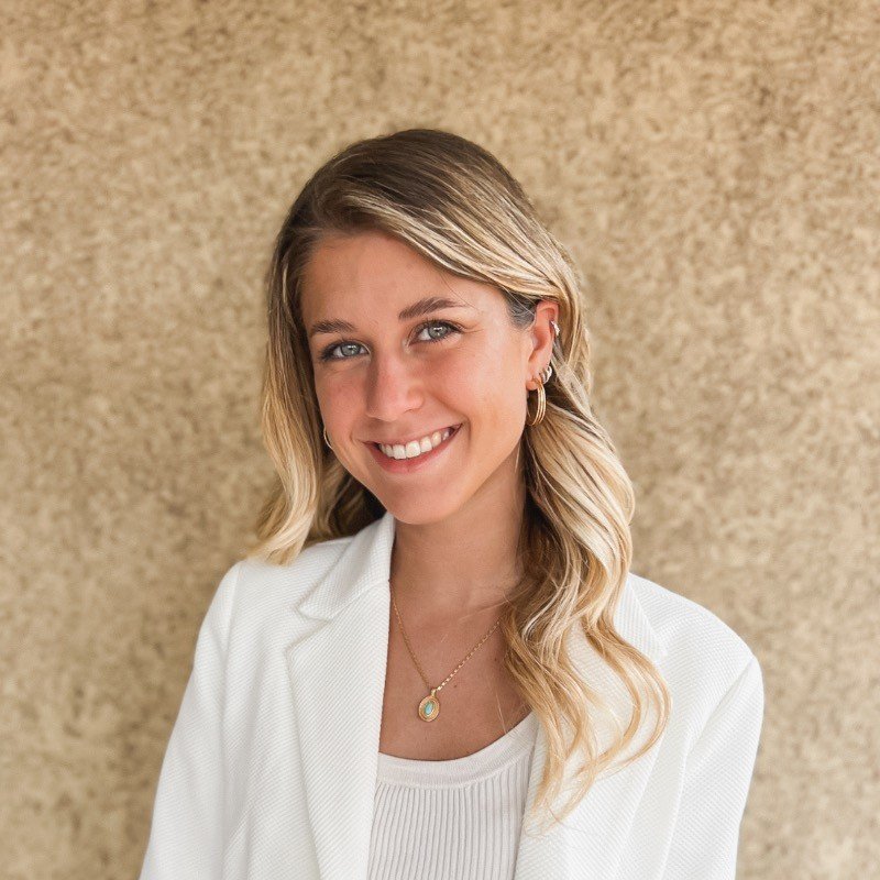 Mujer con cabello rubio y ojos azules, sonriendo, lleva aretes dorados y collar, vestida con blusa blanca y blazer blanco, de fondo beige.