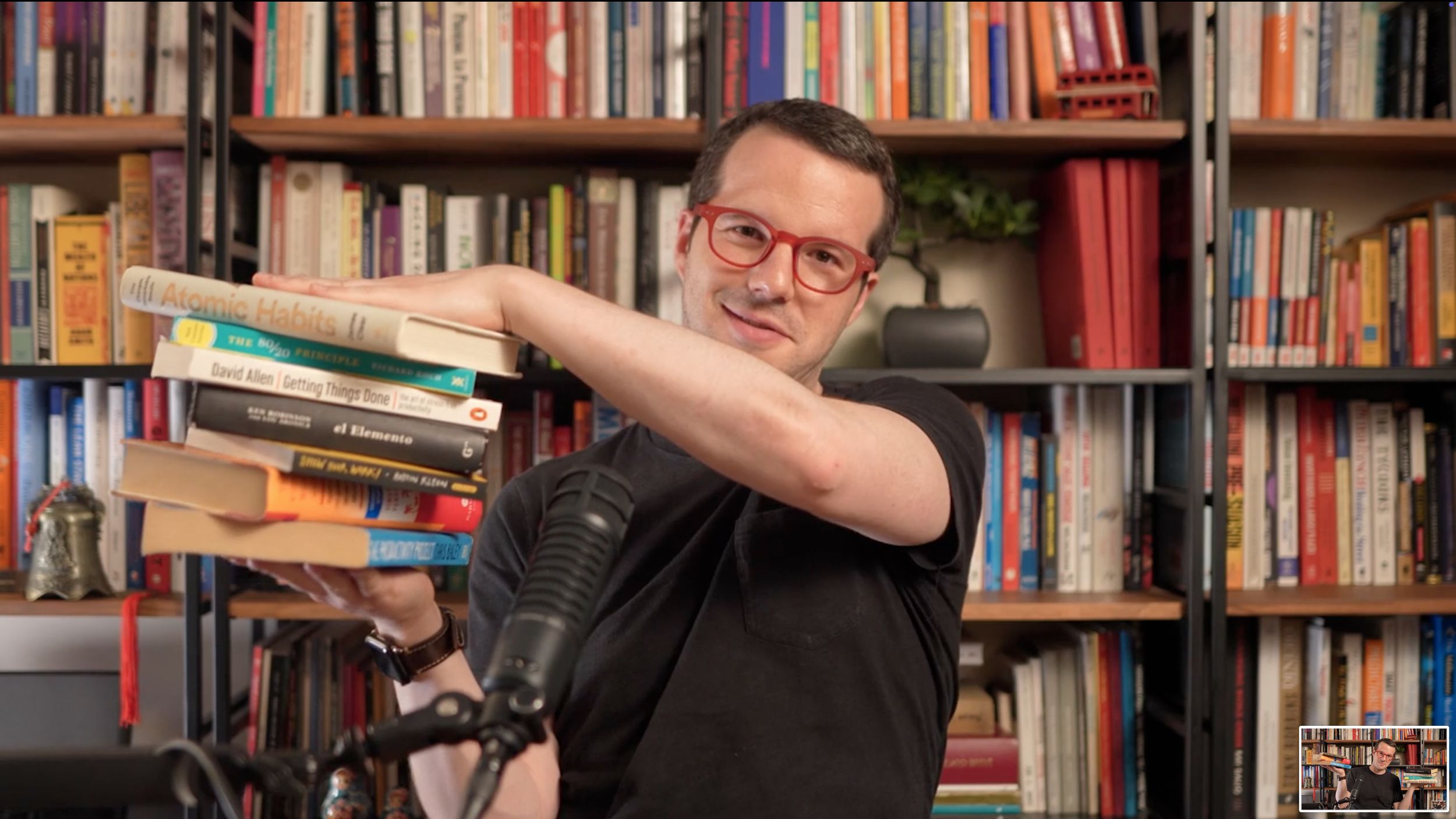 Un hombre con gafas rojas sostiene una pila de libros en una librería o biblioteca, con estanterías llenas de libros al fondo.