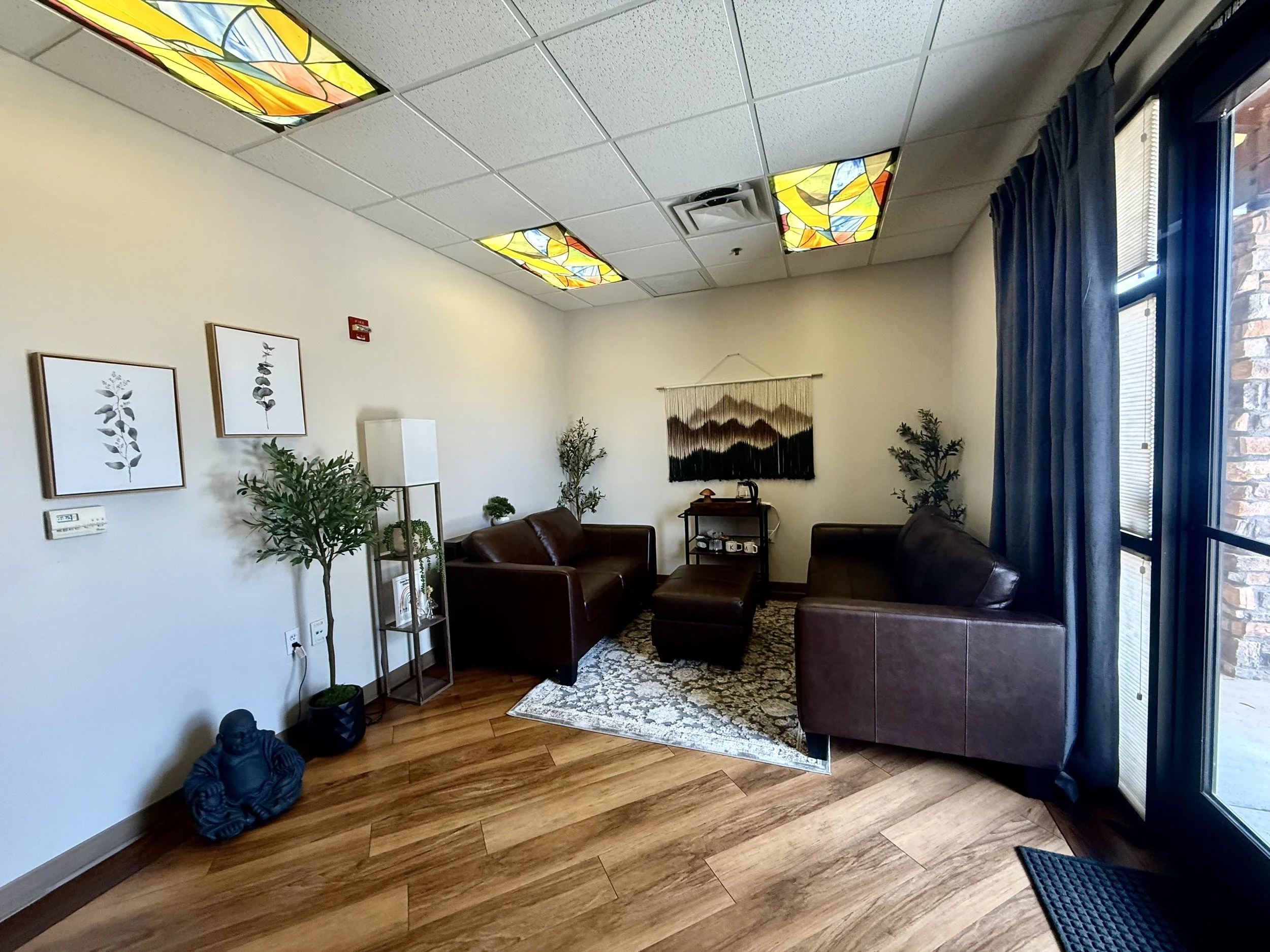 A cozy waiting room with brown leather sofas, framed botanical art on the wall, indoor plants, a patterned rug, and stained glass ceiling lights.