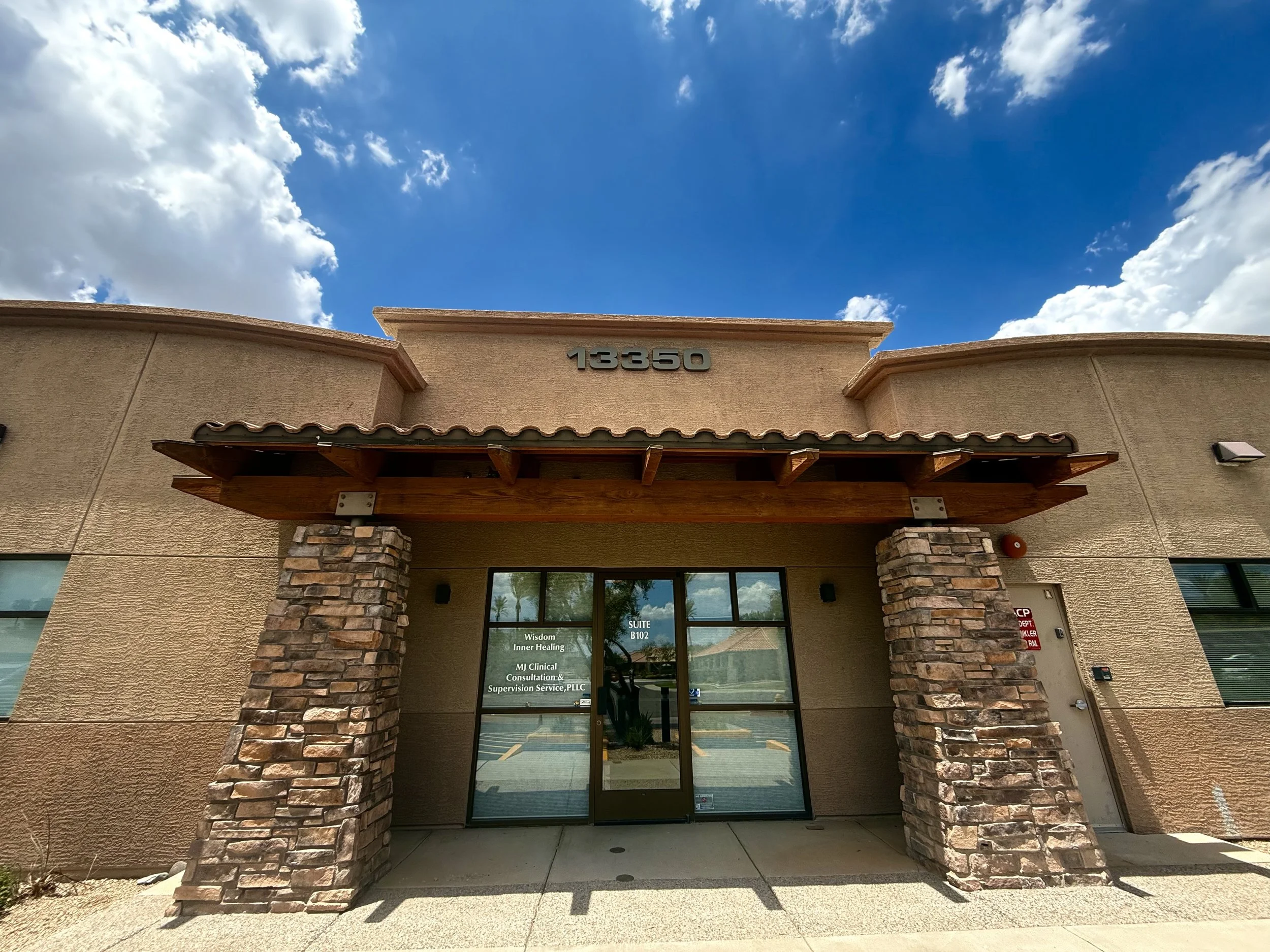 Building with beige stucco walls, stone columns, and a wooden awning, under a blue sky with white clouds.