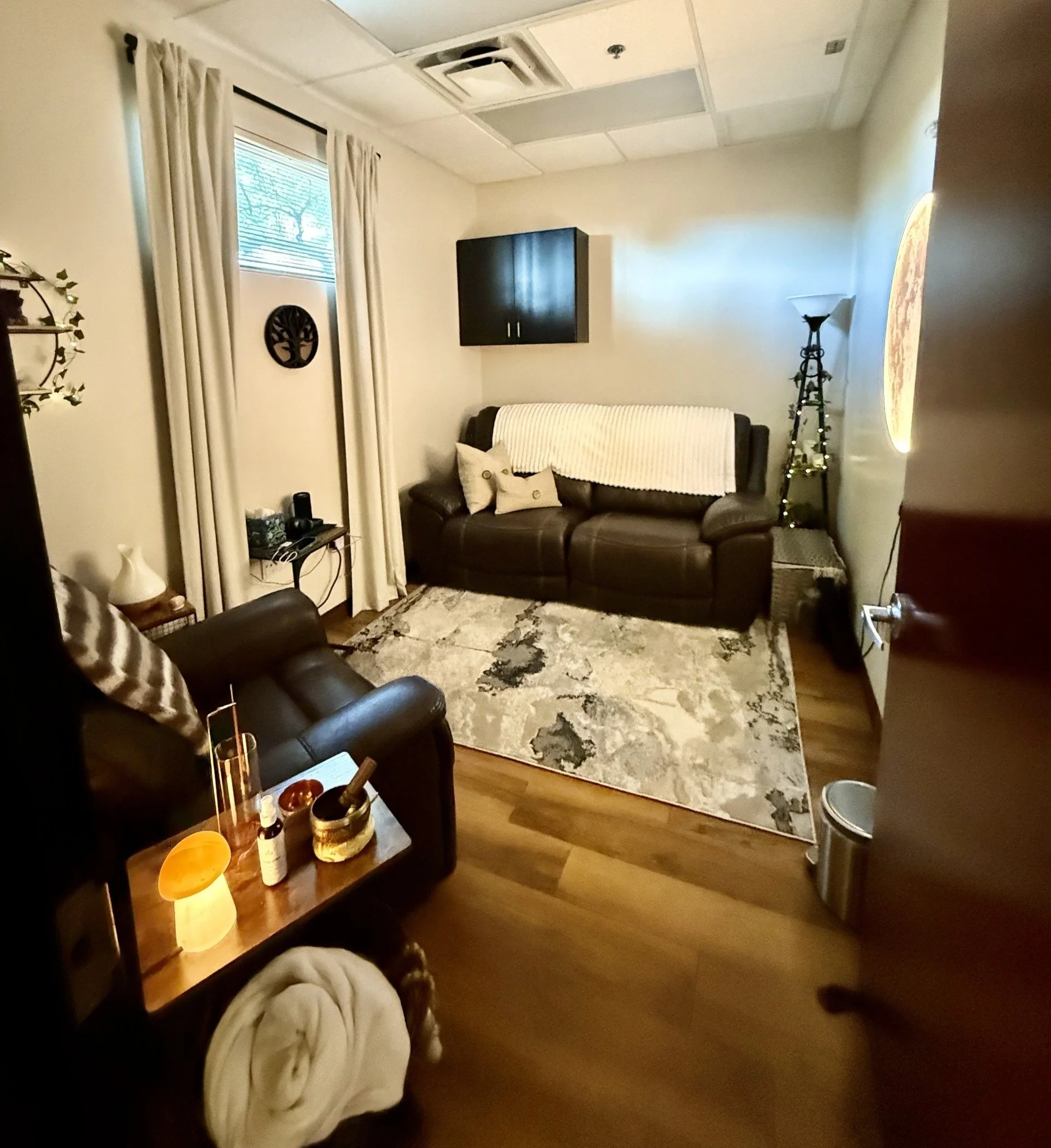 A cozy living room with a brown leather sofa, a white throw blanket, and decorative pillows, a small wooden table with various bottles and a yellow lamp, a patterned area rug, beige walls, white curtains on a window, wall decorations, and a black cabinet mounted on the wall with a clock or artwork beside it.