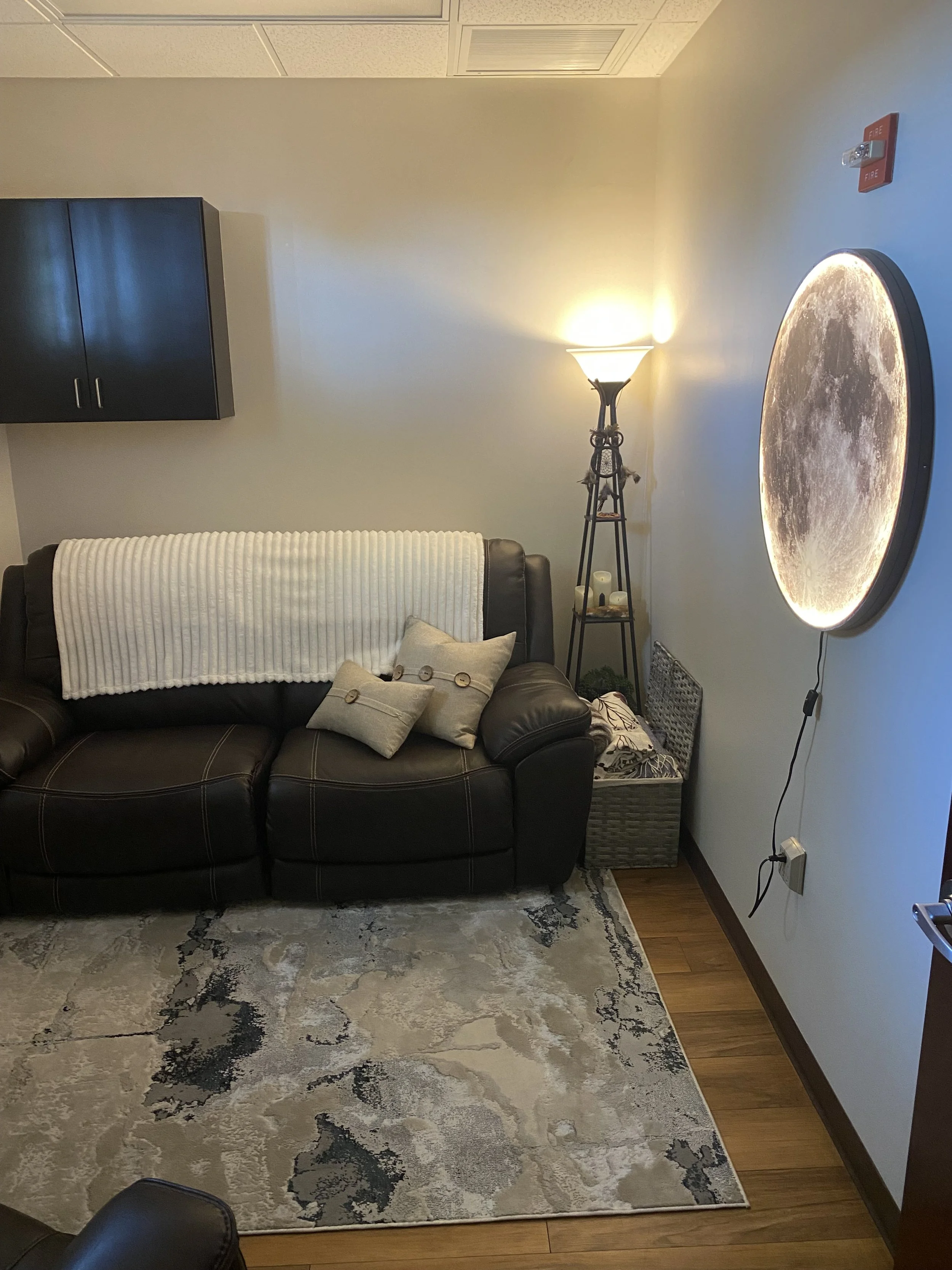 A living room corner with a black leather sofa, a beige throw blanket, and pillows. There is a decorative floor lamp, a small side table with candles, a wall-mounted moon lamp, an area rug, and a wall with a fire alarm and electrical outlets.