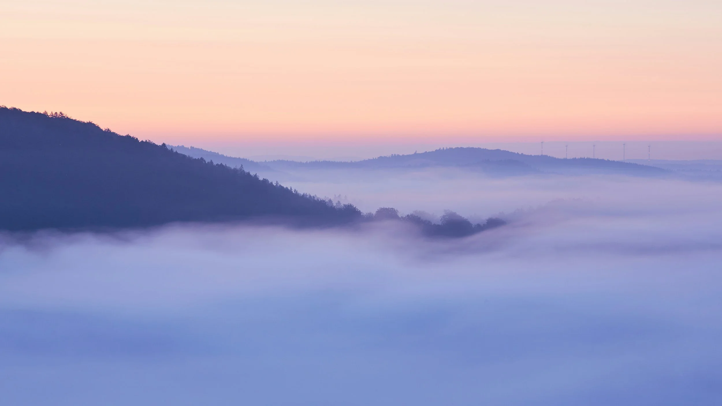 Soft pastel sunrise over rolling hills partially covered in thick fog, with layered ridgelines fading into the distance.