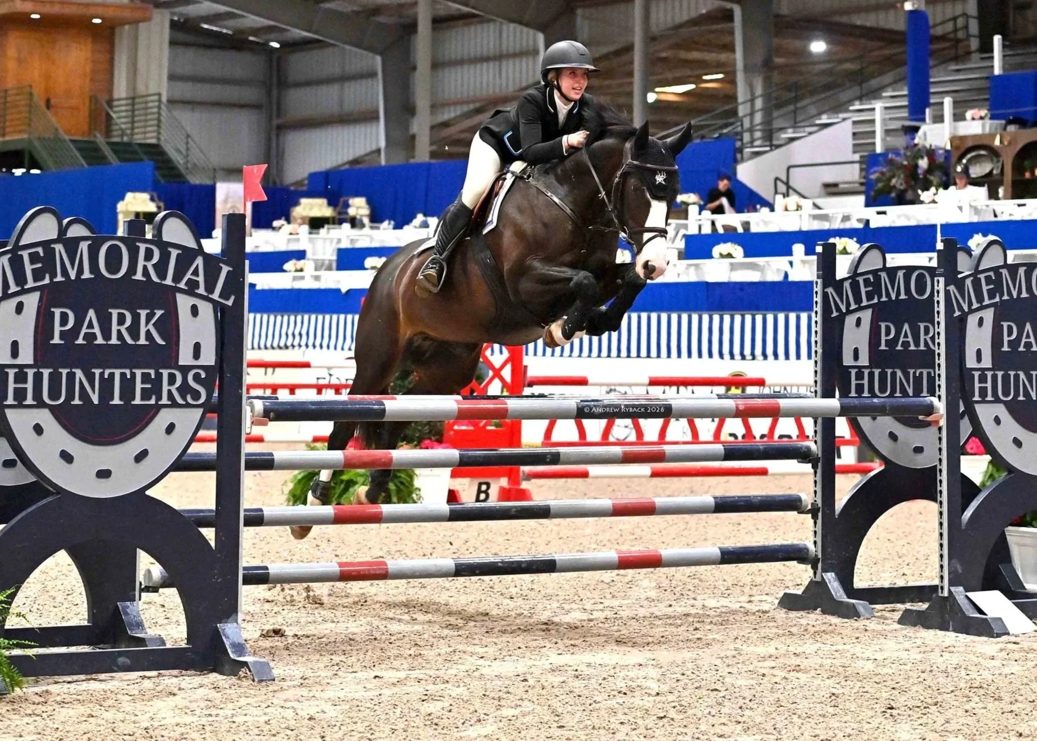 A female equestrian athlete wearing a helmet and riding attire jumps over a hurdle on a brown horse in an indoor equestrian arena decorated with Memorial Park Hunters banners.