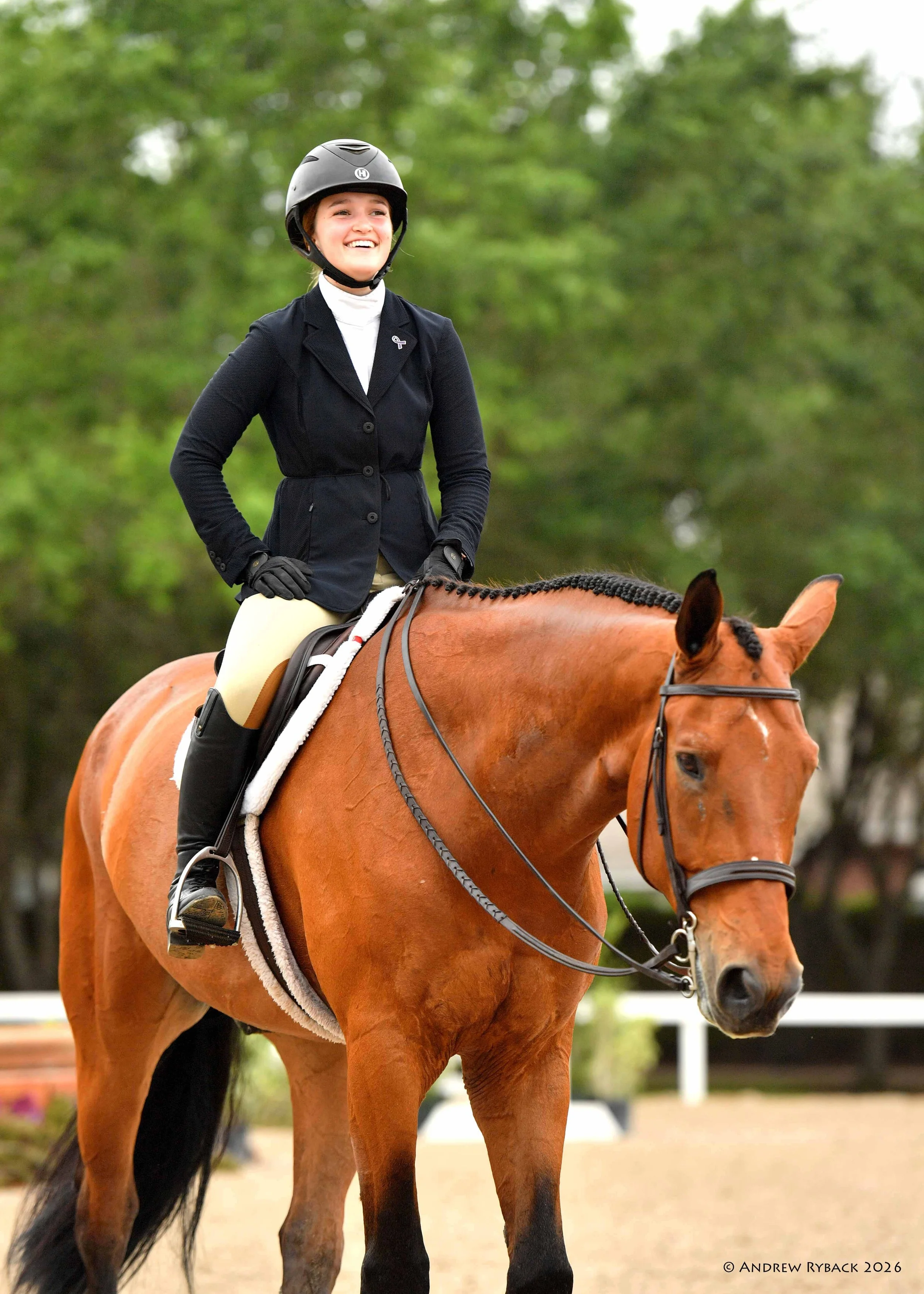 A young woman wearing a black riding helmet, black blazer, and beige riding pants is smiling while riding a chestnut horse with a black mane in an outdoor setting with green trees in the background.